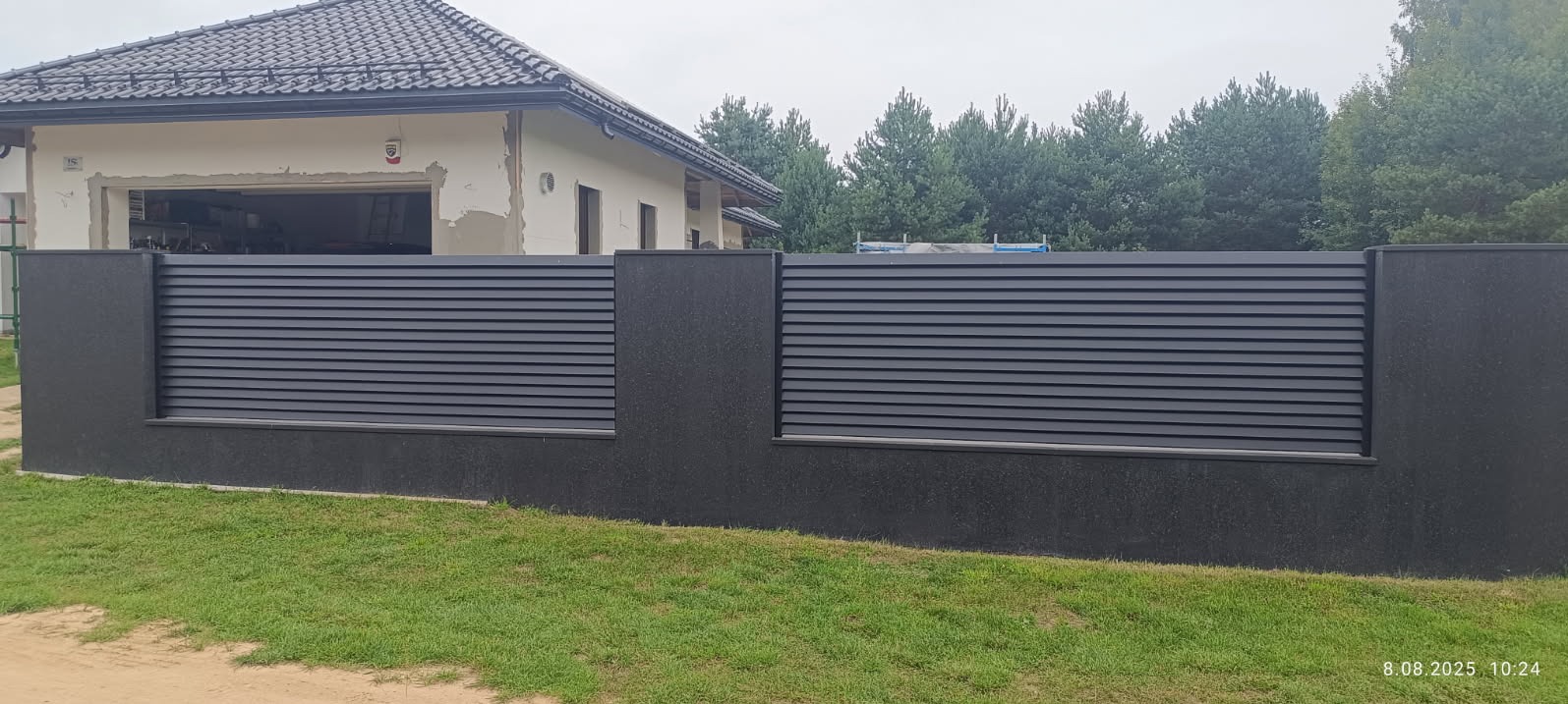 Nowoczesne, grafitowe ogrodzenie panelowe z elementami z fakturą kamienia, na tle domu w trakcie wykończenia i zieleni. Widok z poziomu gruntu.