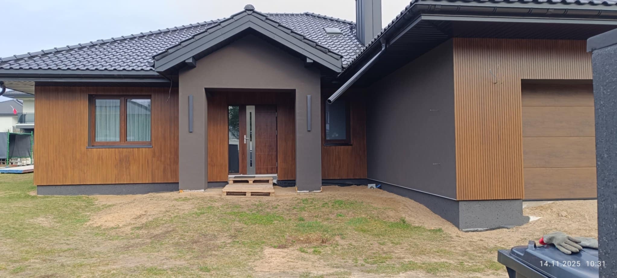 Nowoczesny dom jednorodzinny z elewacją w odcieniach brązu i szarości, z widocznymi panelami elewacyjnymi i ciemną dachówką. Wejście z drewnianymi schodami.