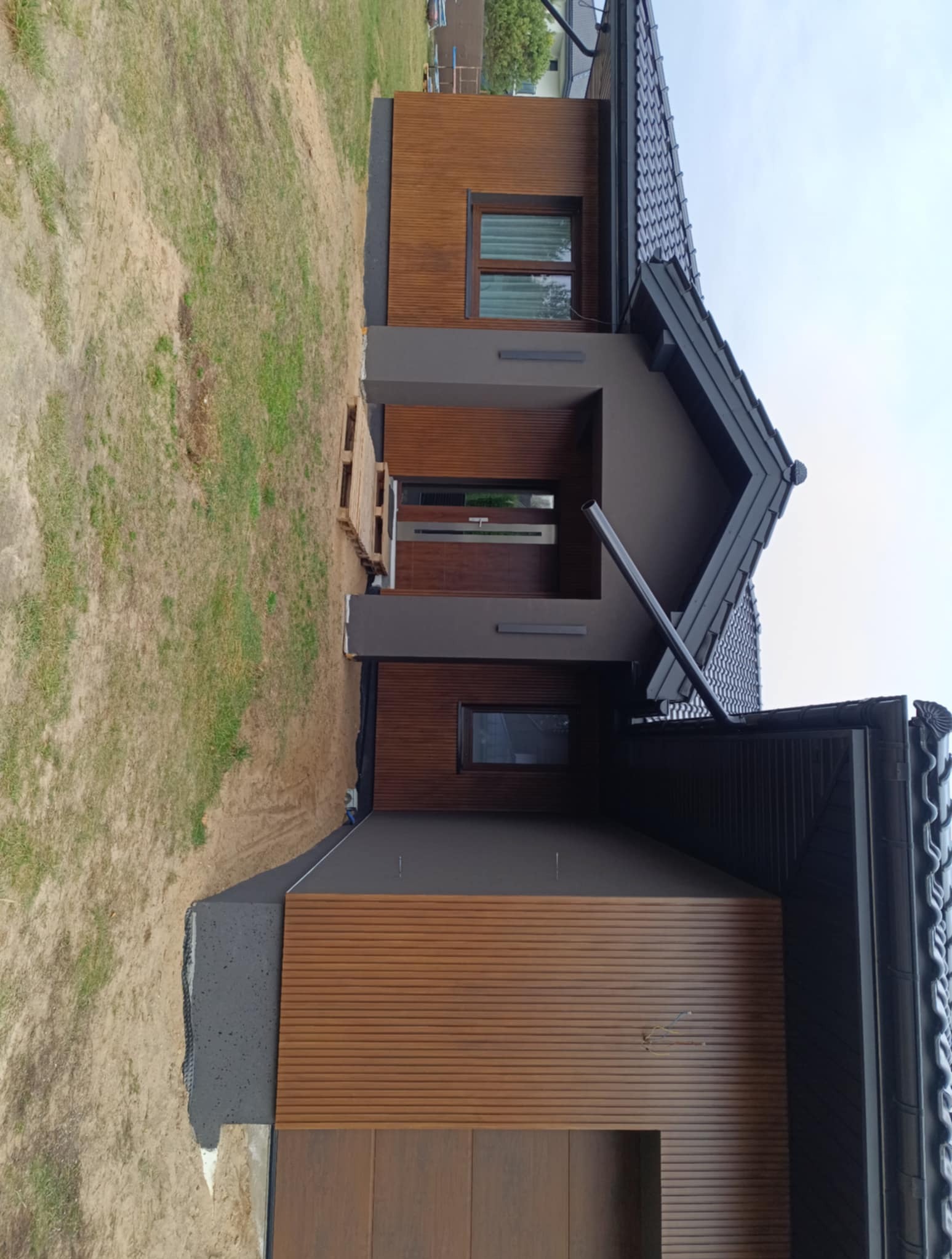 Nowoczesny dom z elewacją z paneli drewnopodobnych i szarą stolarką okienną. Widoczne detale wykończenia i elementy drenażu. Ujęcie z poziomu gruntu.