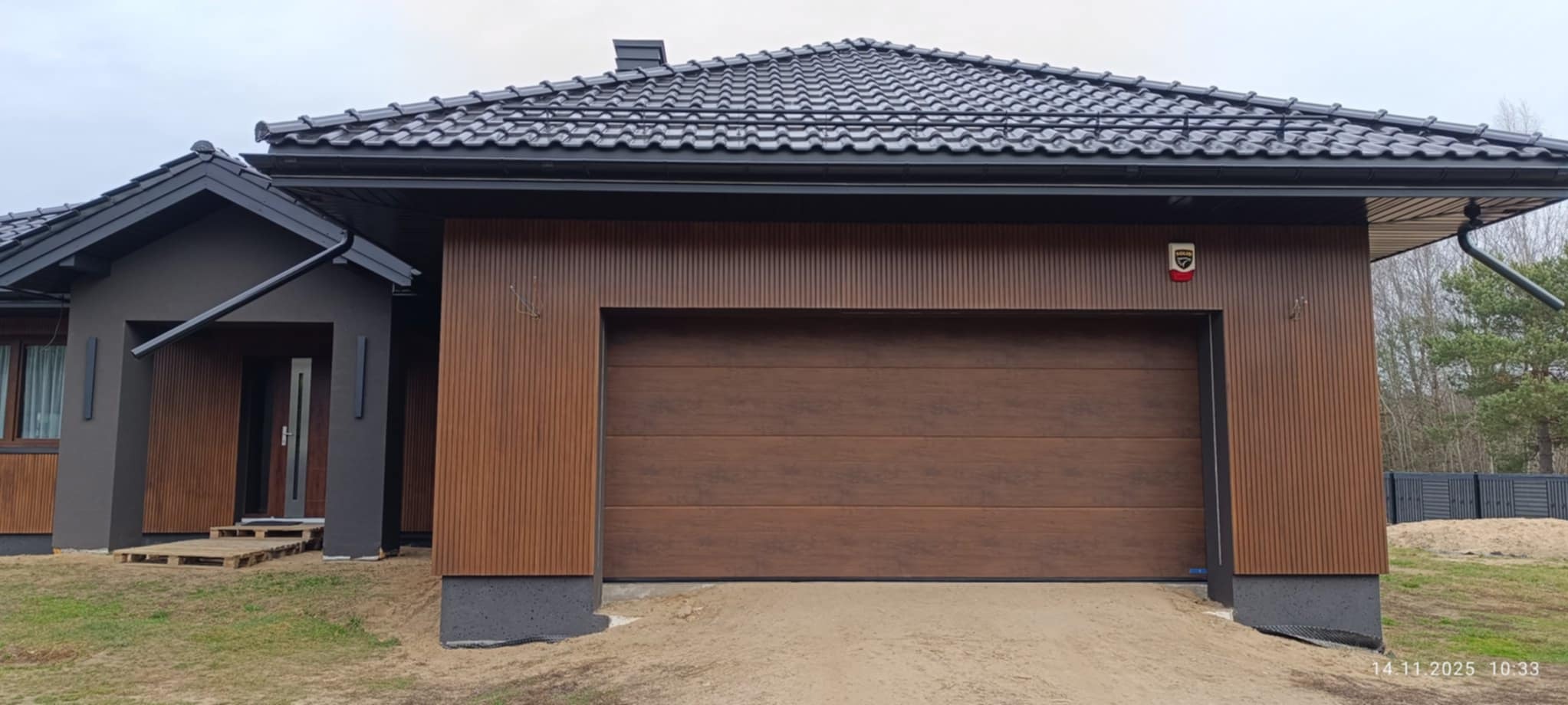 Nowoczesny dom z garażem w brązowej okładzinie elewacyjnej. Ciemna dachówka i grafitowe elementy wykończeniowe. Widoczny system alarmowy.