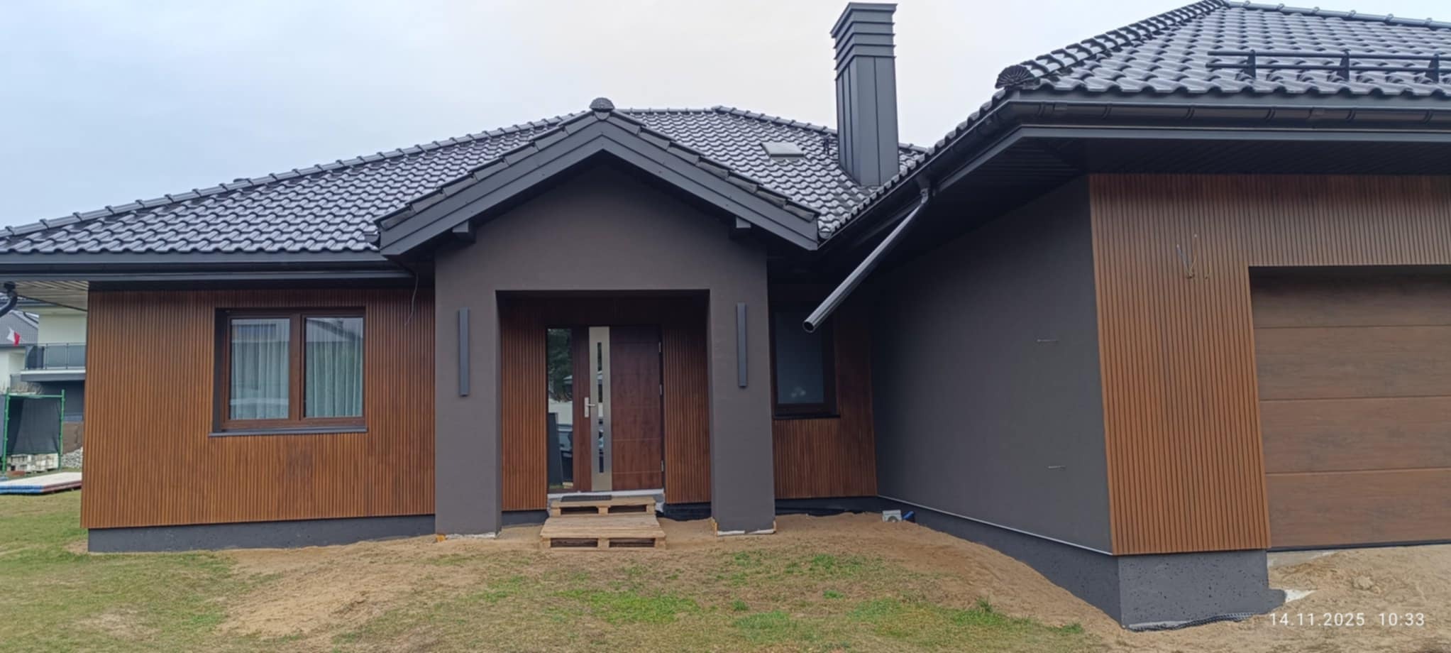 Nowoczesny dom jednorodzinny z elewacją w odcieniach brązu i szarości, z widocznym wejściem i garażem. Dach pokryty grafitową dachówką. Ujęcie z poziomu gruntu.