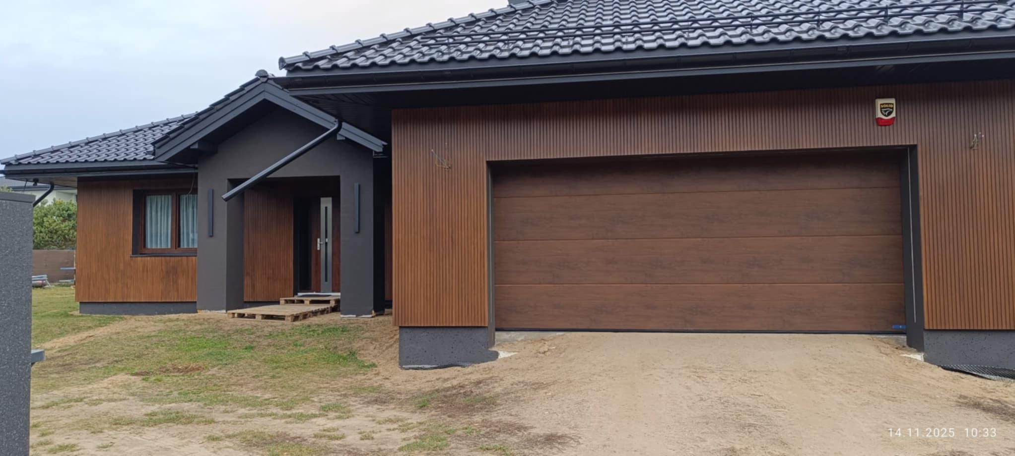 Nowoczesny dom jednorodzinny z brązową elewacją imitującą drewno i antracytową stolarką okienną oraz dachem. Widoczny garaż z bramą segmentową.