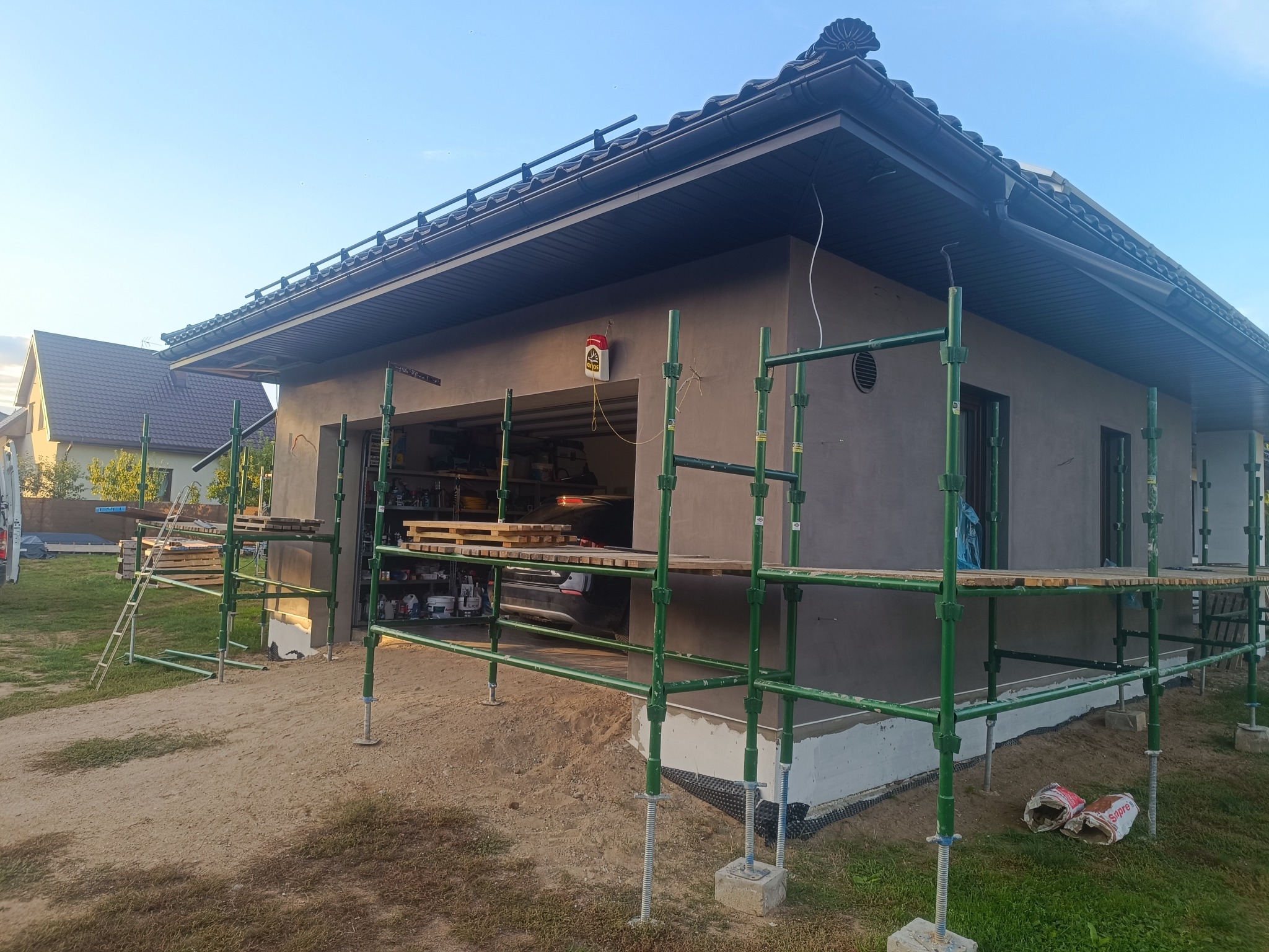 Dom jednorodzinny w trakcie prac elewacyjnych, otoczony rusztowaniem. Ściany pokryte szarą warstwą, widoczne materiały budowlane i narzędzia. Ujęcie z zewnątrz, w dziennym świetle.