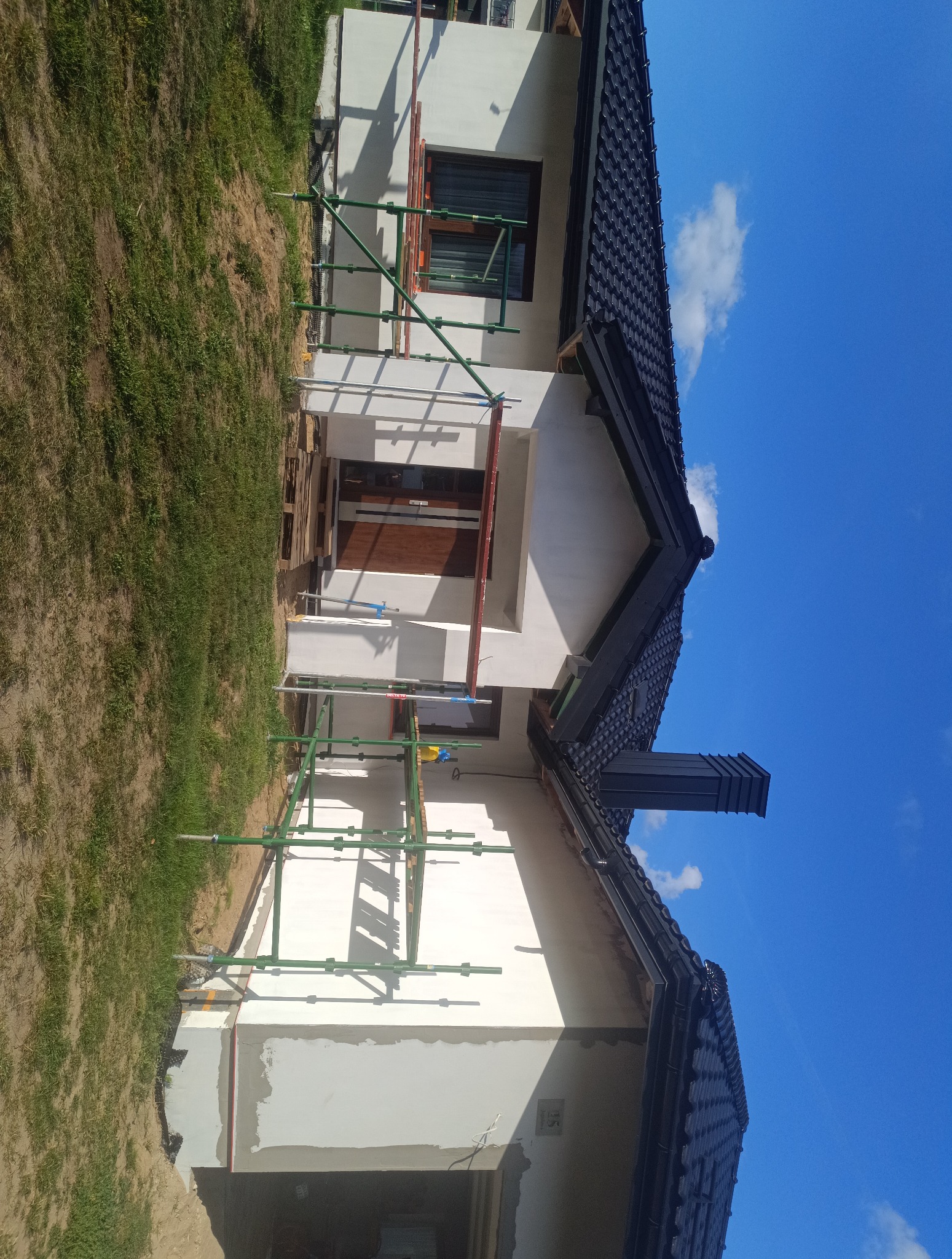 Dom jednorodzinny w trakcie prac elewacyjnych. Widoczne rusztowania, fragment otynkowanej ściany i ciemna dachówka. Błękitne niebo w tle.