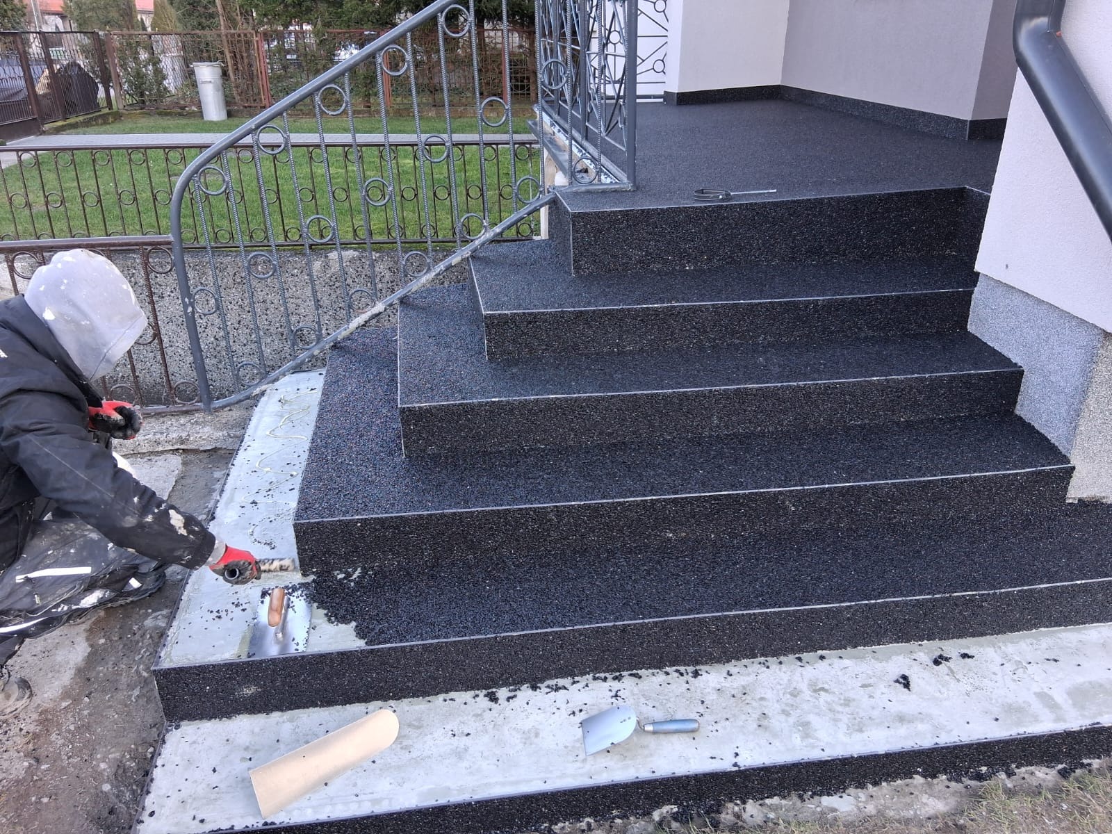 Aplikacja czarnej, drobnoziarnistej nawierzchni na schodach wejściowych. Pracownik w kombinezonie rozprowadza masę szpachelką. Widoczne narzędzia i fragment balustrady.