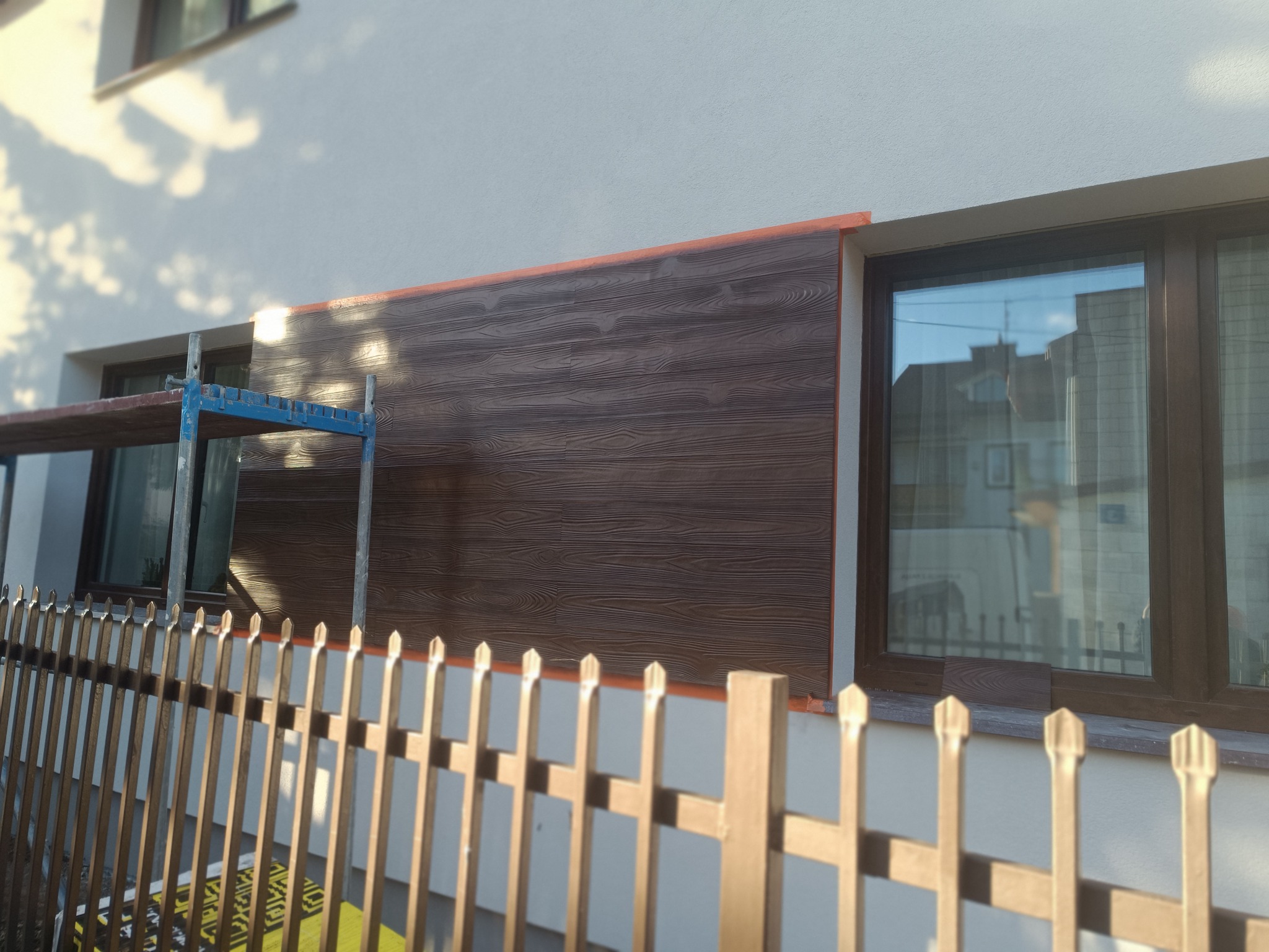 Fragment elewacji budynku z brązową okładziną imitującą drewno, widoczne rusztowanie i ogrodzenie. Częściowo zakończony montaż paneli elewacyjnych.