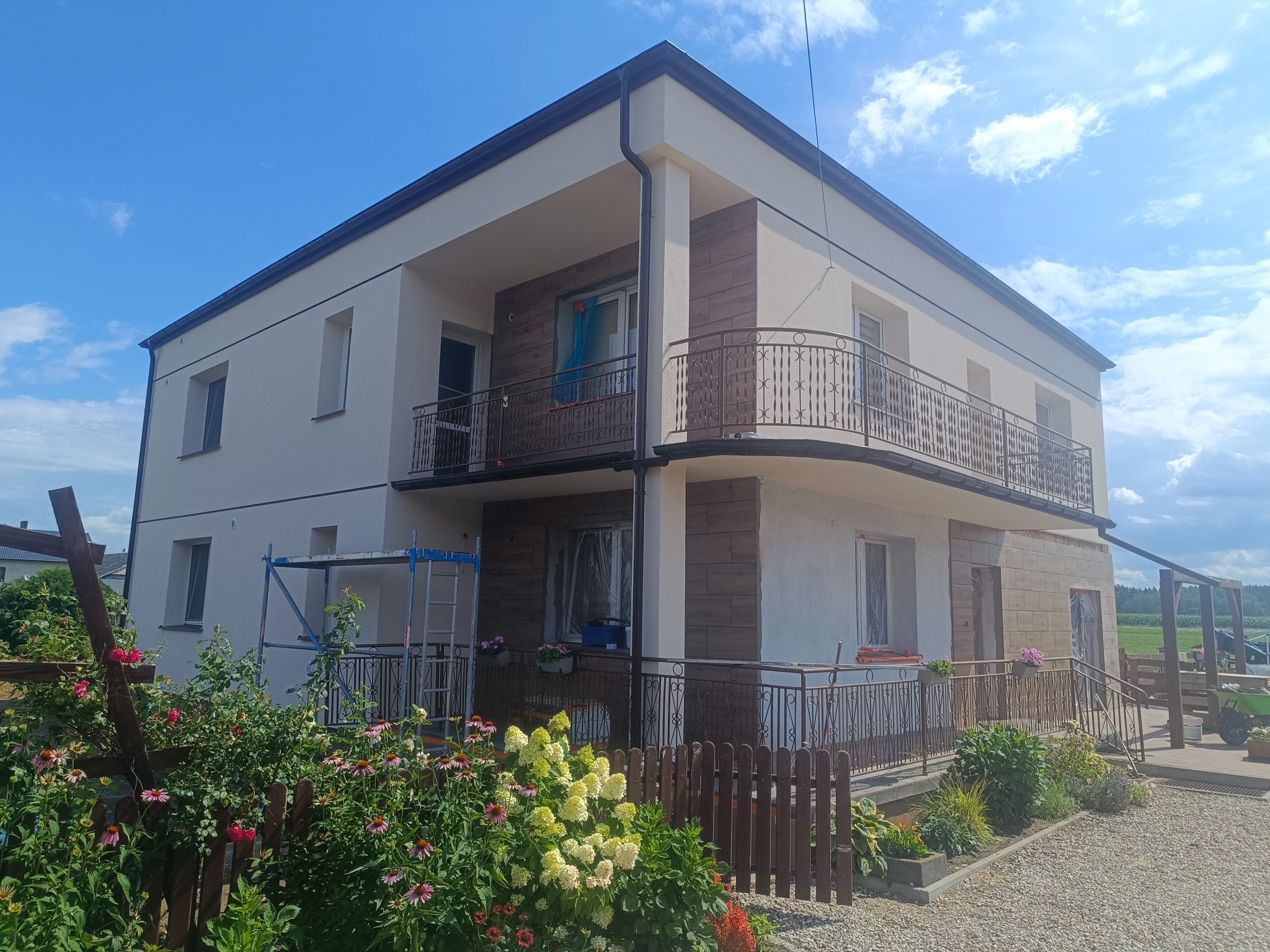 Dwukondygnacyjny dom z elewacją w trakcie prac wykończeniowych. Część ścian pomalowana na biało, część wyłożona płytkami imitującymi drewno. Widoczne rusztowania i zabezpieczenia okien.