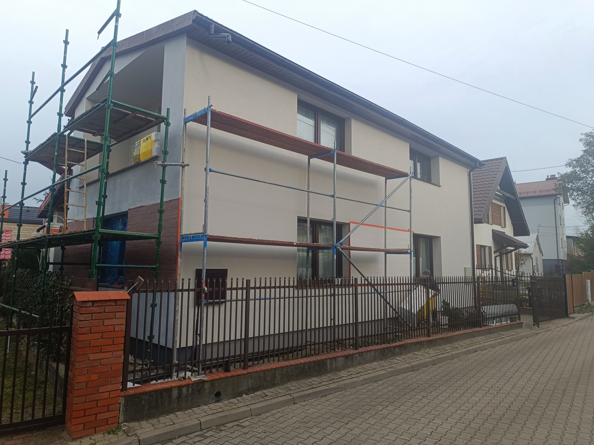 Dwukondygnacyjny dom w trakcie ocieplania elewacji, widoczne rusztowania i częściowo ocieplona ściana z imitacją drewna. Prace w toku, pochmurny dzień.