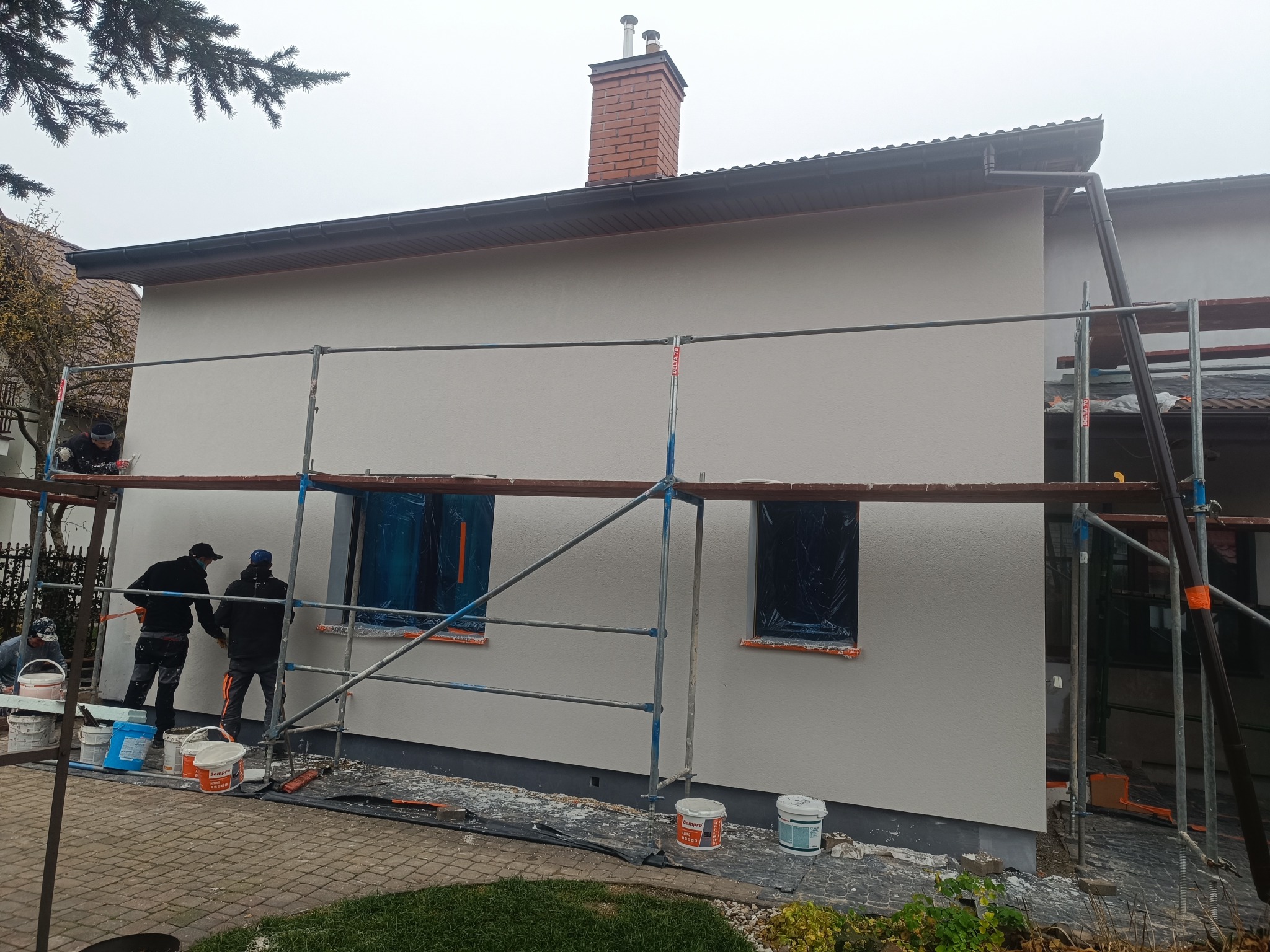 Prace na elewacji domu jednorodzinnego. Ekipa na rusztowaniu, zabezpieczone okna folią, widoczne wiadra z farbą. Świeżo nałożona, jasna warstwa tynku.