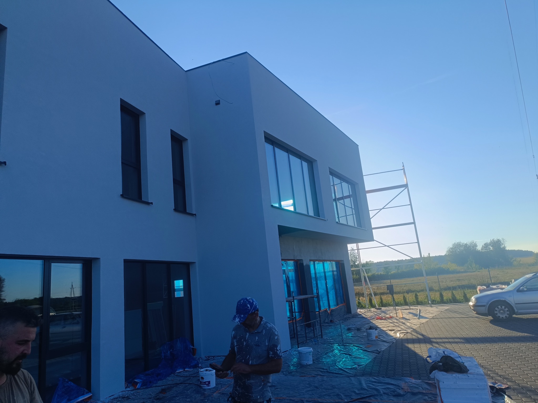 Nowoczesny dom jednorodzinny po malowaniu elewacji. Widoczne rusztowania, zabezpieczenia okien folią i pracownik z farbą. Jasna kolorystyka budynku.