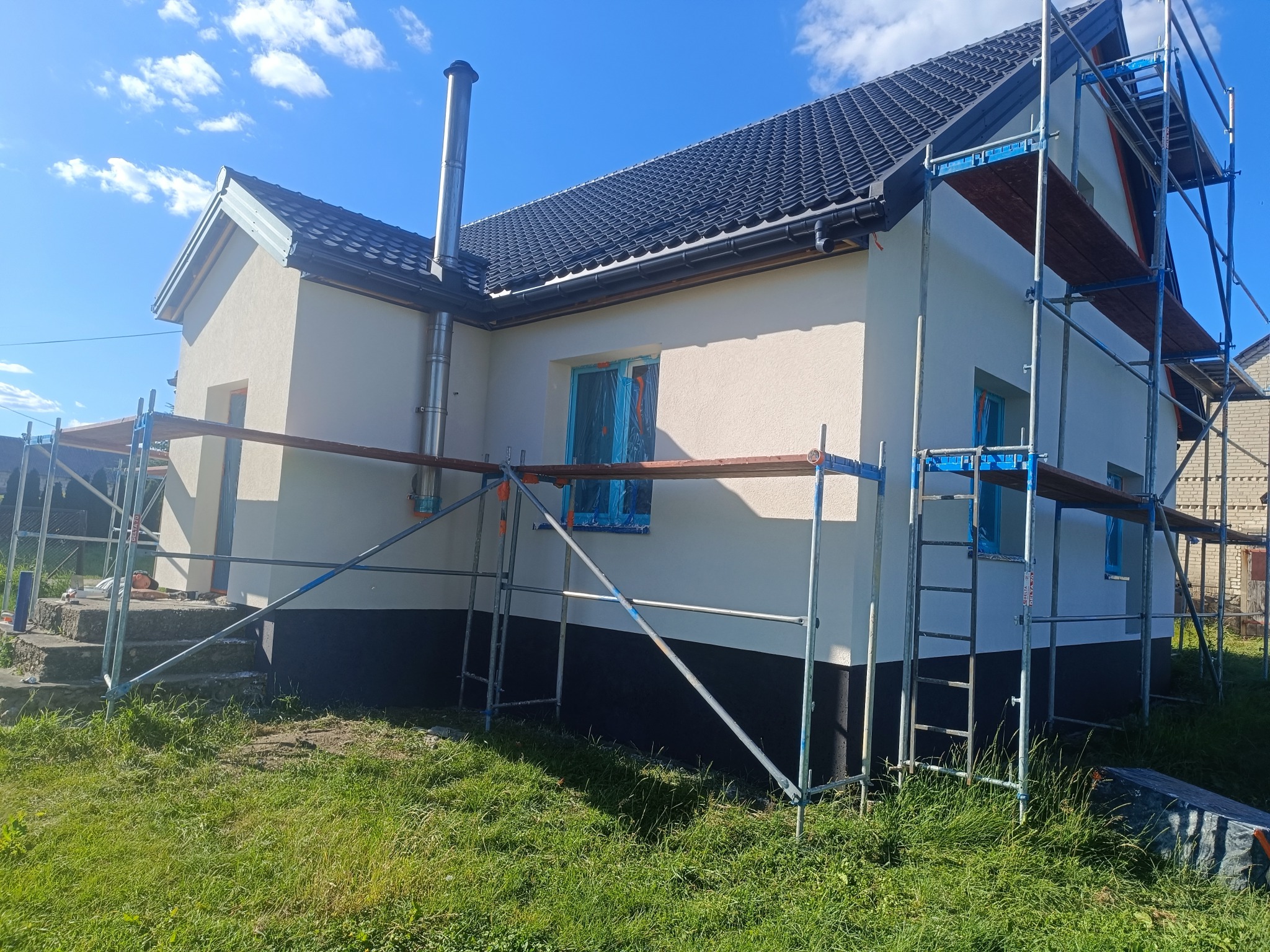 Dom jednorodzinny z rusztowaniem, po nałożeniu jasnego tynku. Okna zabezpieczone folią. Widoczny fragment dachu z kominem i trawnik.