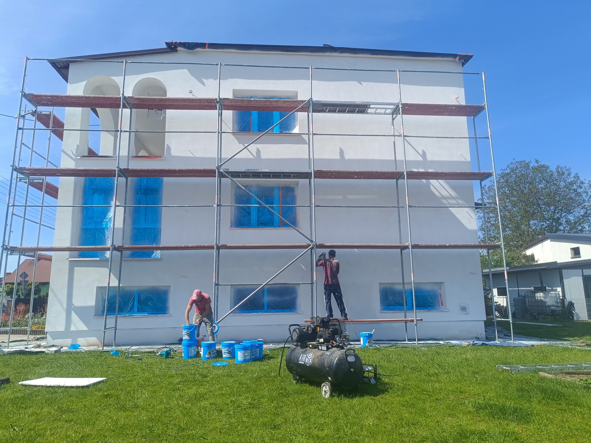 Dwóch pracowników na rusztowaniu przy białej elewacji domu. Okna zabezpieczone folią, widoczne wiadra z farbą i kompresor. Prace w toku, słoneczny dzień.