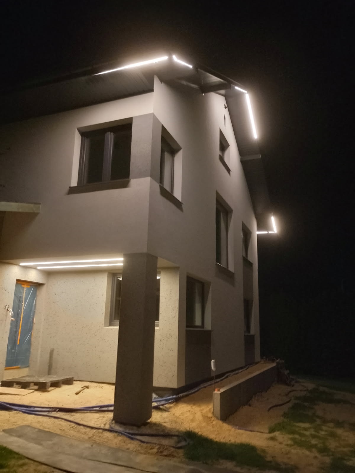 Oświetlona elewacja domu w nocy. Nowoczesne lampy LED podkreślają architekturę budynku. Widoczne okna i fragment terenu wokół domu.