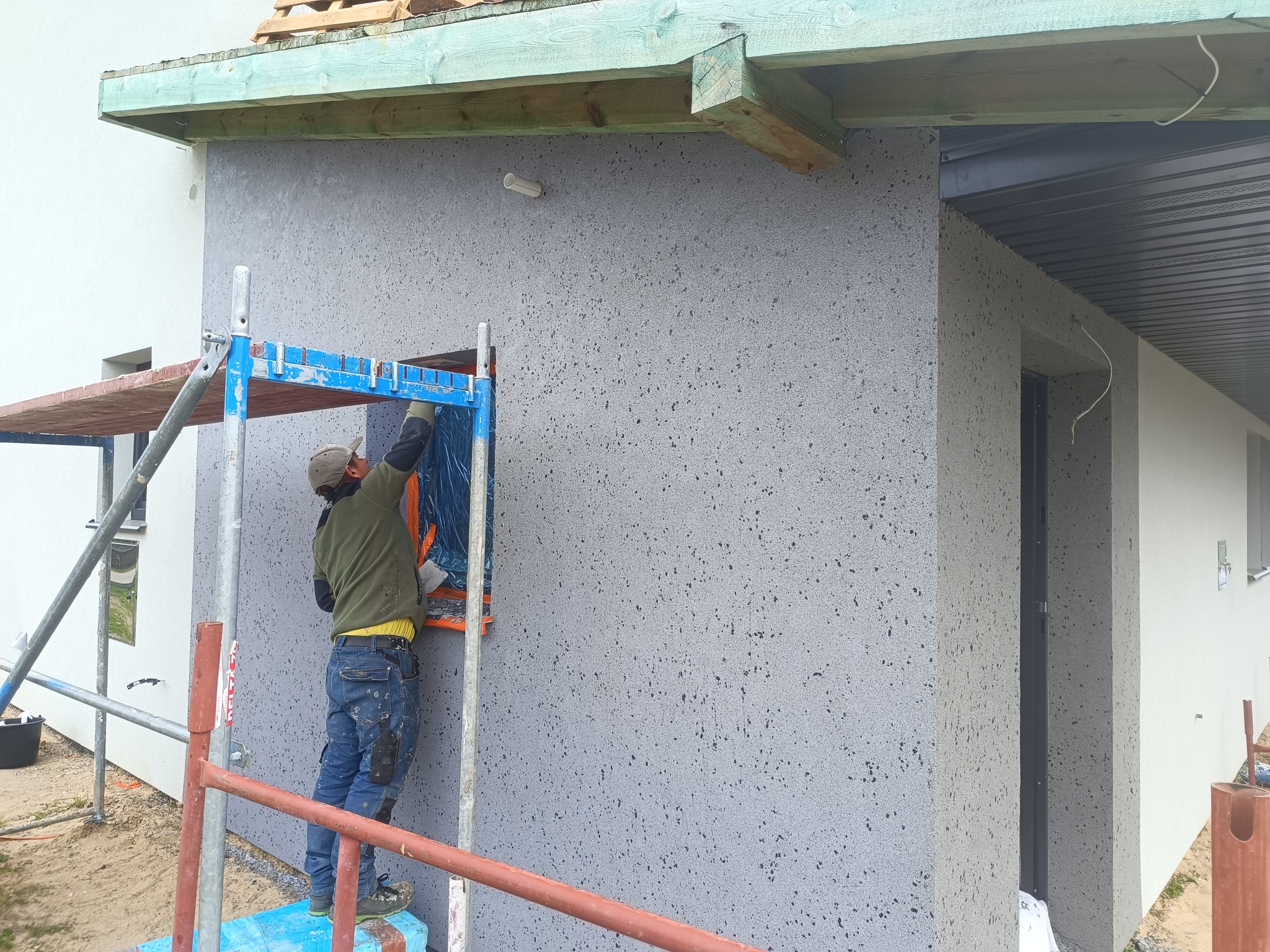 Pracownik na rusztowaniu zabezpiecza okno folią przed nałożeniem szarego tynku barankowego na elewacji domu. Widoczne drewniane elementy konstrukcyjne dachu.