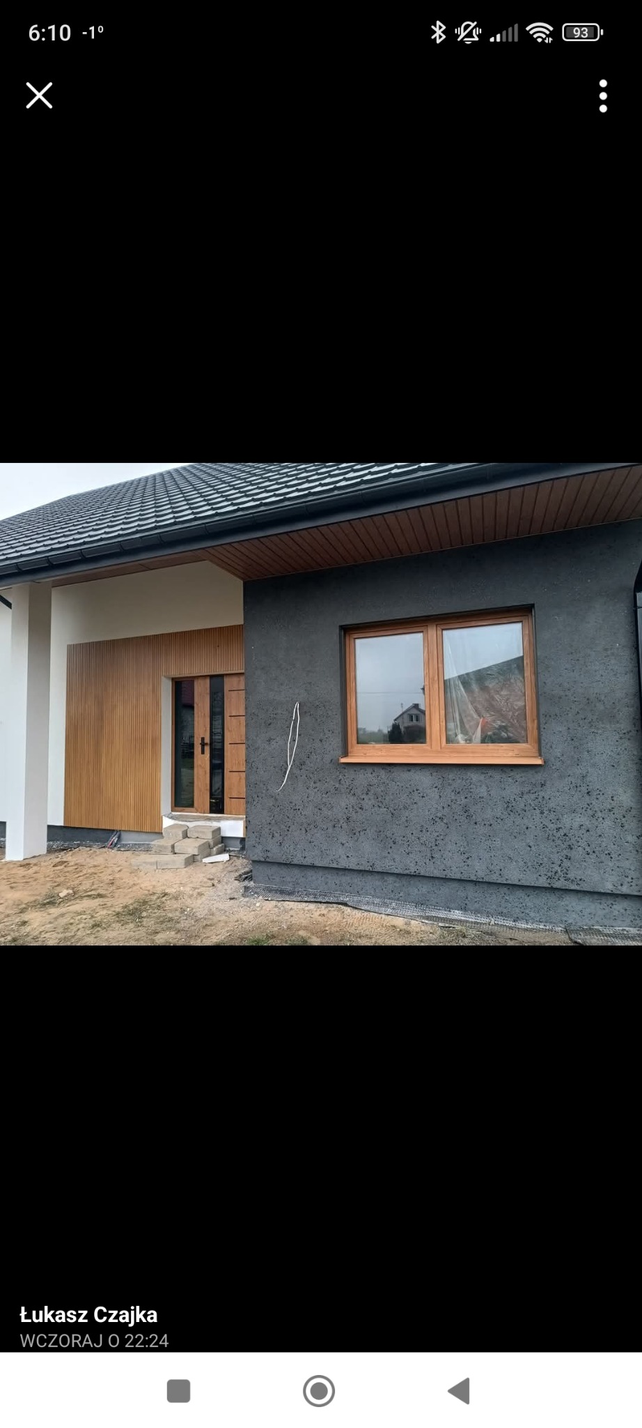 Dom jednorodzinny z elewacją w odcieniach szarości i drewna. Widoczne okno z drewnianą ramą i fragment schodów wejściowych. Nowoczesny styl.