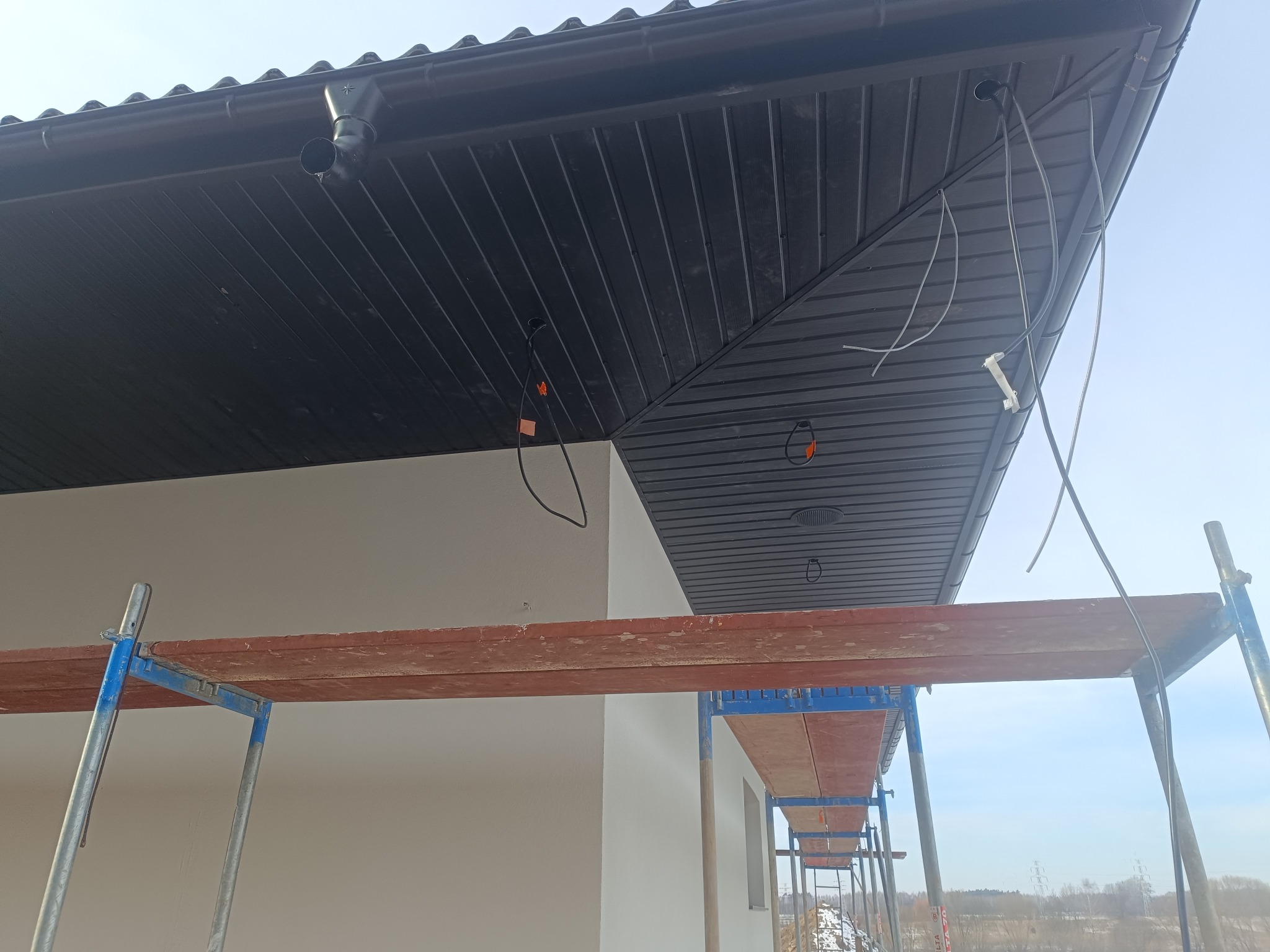 Montaż podbitki dachowej z widocznymi przewodami i rusztowaniem. Ujęcie z dołu, jasne niebo w tle. Widoczne elementy systemu rynnowego.