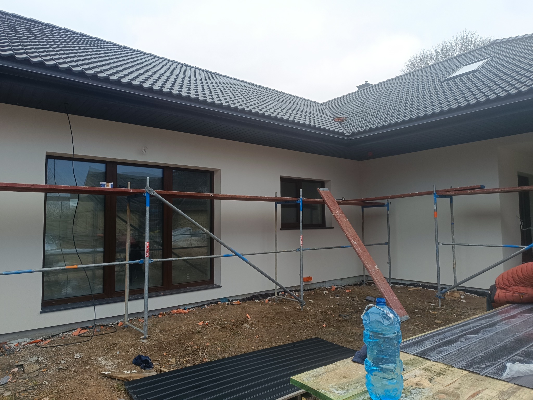 Dom jednorodzinny po nałożeniu jasnej elewacji, widoczne rusztowania i butelka wody. Nowoczesna architektura z ciemną dachówką.