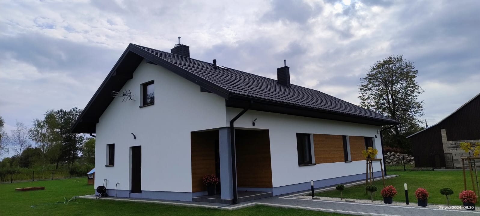 Biały dom jednorodzinny z ciemnym dachem i elementami wykończenia w drewnie, otoczony zielonym trawnikiem i ozdobnymi roślinami. Widok z boku, pochmurne niebo.