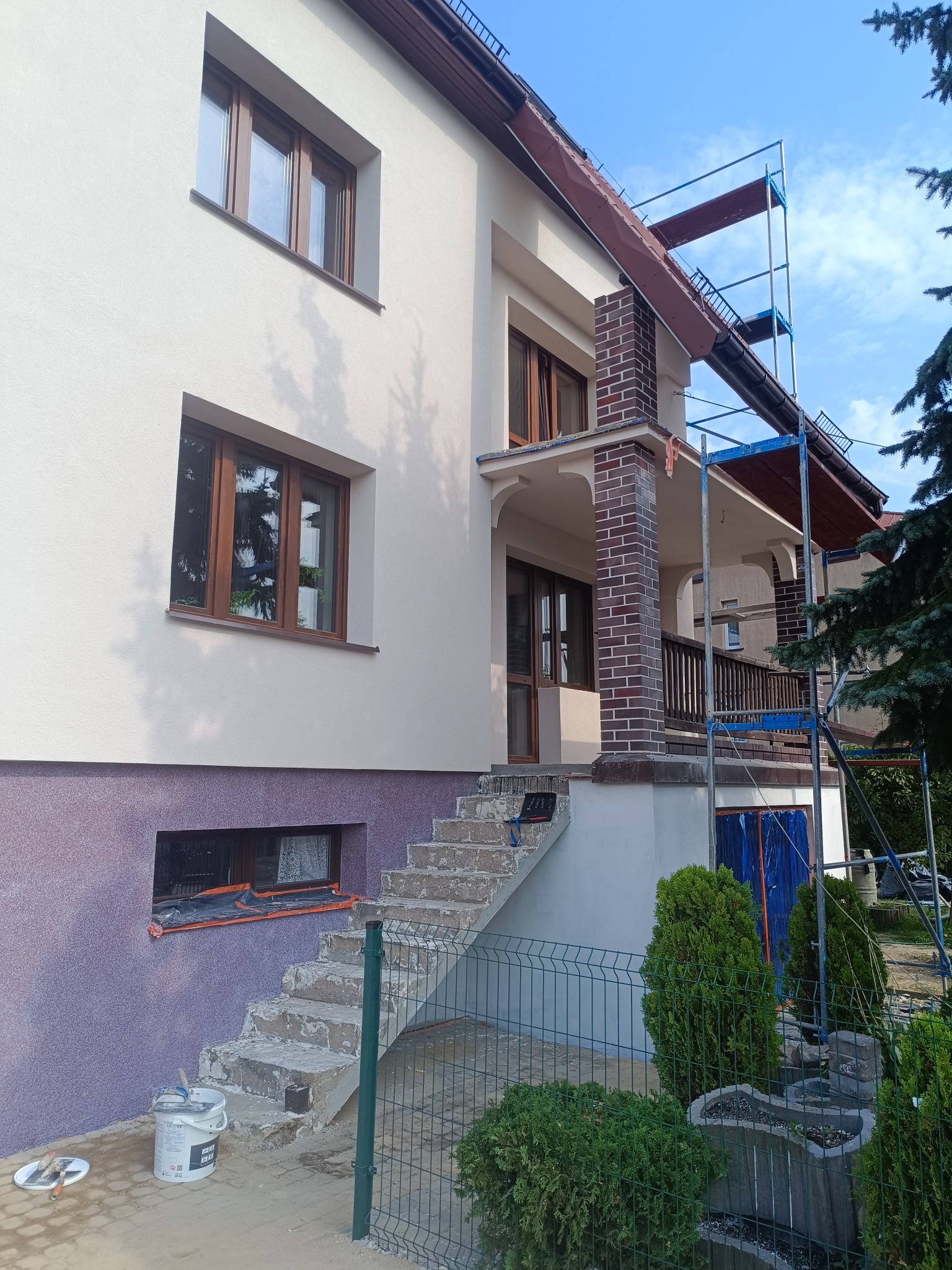 Dom po renowacji elewacji: kremowa góra, fioletowy dół. Widoczne schody wejściowe, rusztowanie i wiadro z farbą. Brązowe okna i ceglane kolumny werandy.