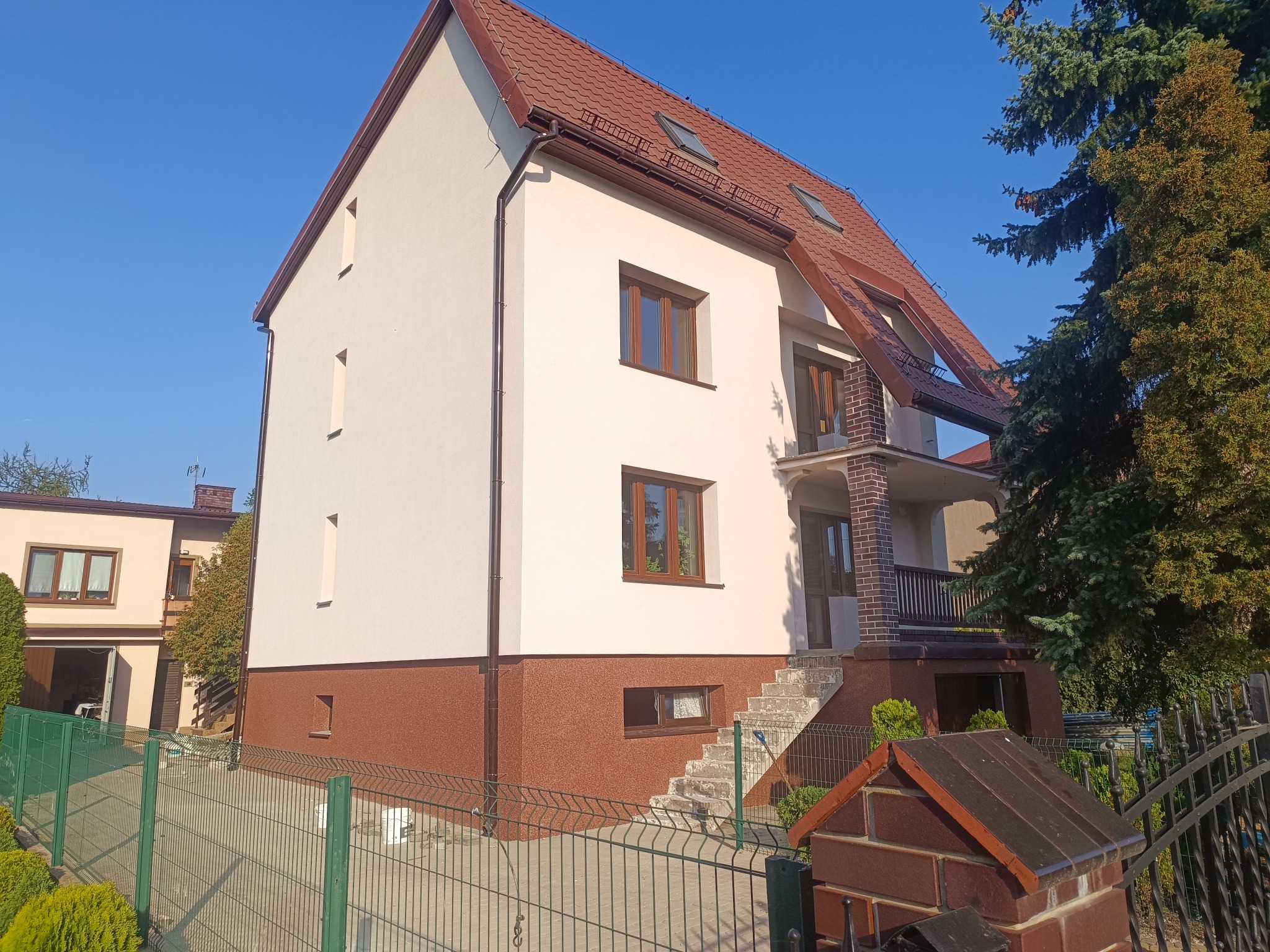 Dwukondygnacyjny dom z beżową elewacją i brązowym cokołem, widoczny z perspektywy ogrodu. Czerwony dach z oknami połaciowymi, brązowe rynny i okna.