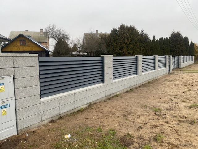 Nowoczesne ogrodzenie posesji z szarymi, poziomymi panelami i kamiennymi słupkami, widok z zewnątrz na tle zabudowy jednorodzinnej i drzew.