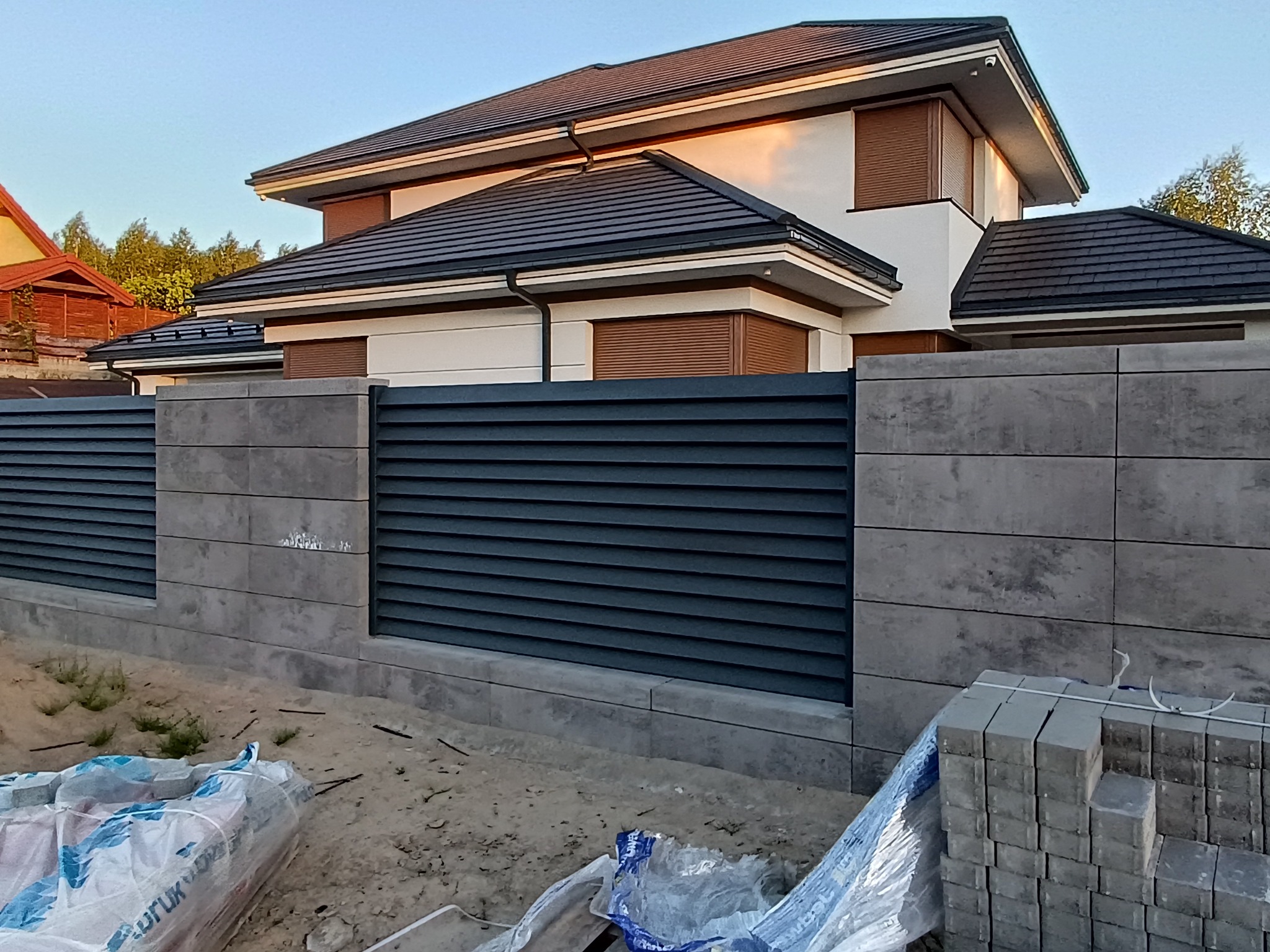 Nowoczesny dom z szarą, lamelową bramą i ogrodzeniem z betonowych bloczków. Na pierwszym planie materiały budowlane.