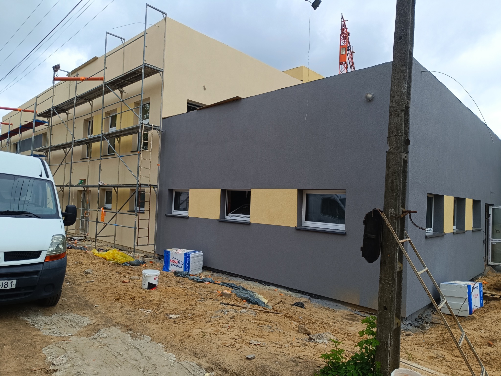 Budynek po nałożeniu szarego tynku z żółtymi akcentami pod oknami. Część budynku w trakcie prac, widoczne rusztowania i materiały budowlane.