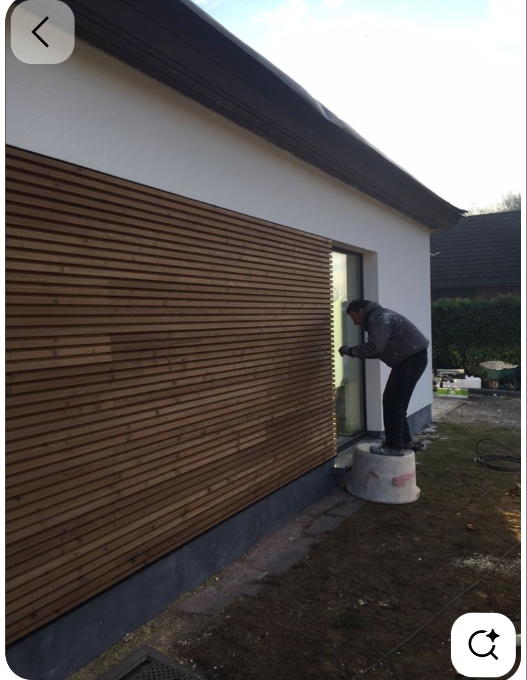 Pracownik na podeście zabezpiecza okno taśmą przed malowaniem elewacji domu z drewnianą okładziną i białą górną częścią. Widoczne narzędzia i otoczenie.