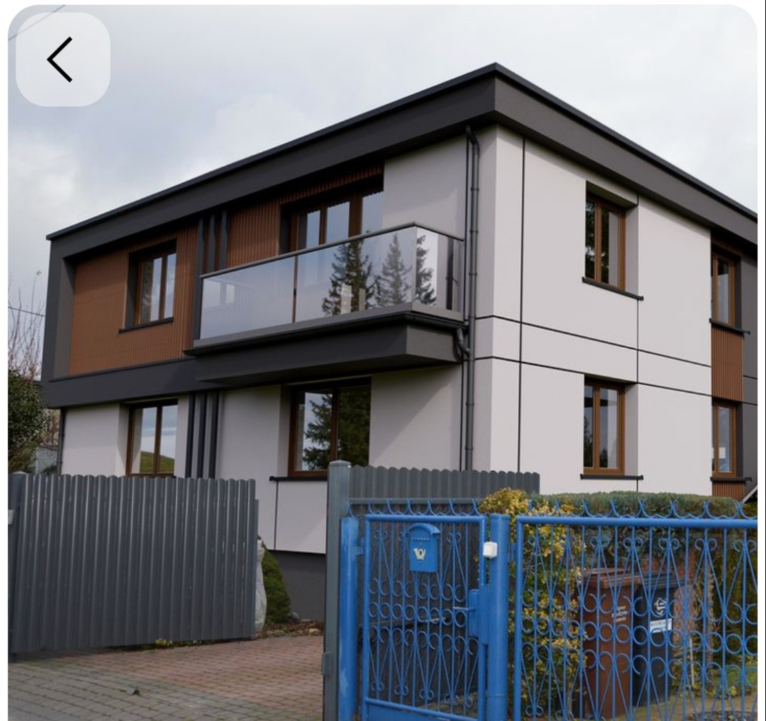 Dwukondygnacyjny dom z balkonem i nowoczesną elewacją w odcieniach bieli, brązu i szarości, widziany zza niebieskiego, metalowego ogrodzenia z bramą.