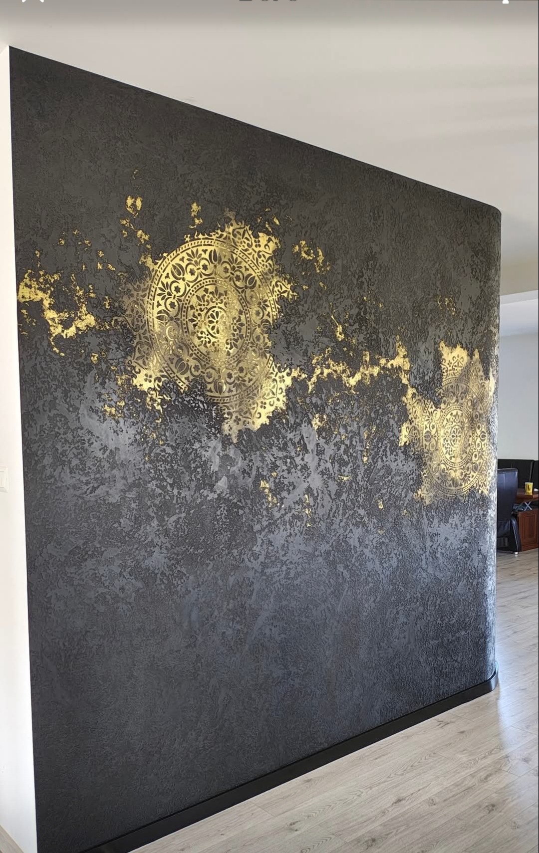 Dekoracyjna ściana w odcieniach szarości z efektem przetarć i złotymi, okrągłymi ornamentami. Widoczna faktura ściany i subtelne przejścia kolorystyczne.