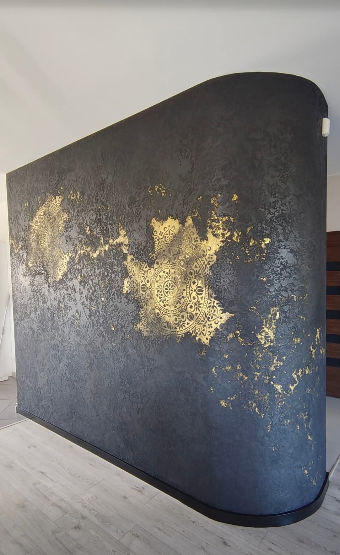 Ciemna ściana z dekoracyjną, złotą strukturą i ornamentami w kształcie mandali. Wykończenie wnętrza z efektem artystycznym, widoczne detale faktury i załamania światła.