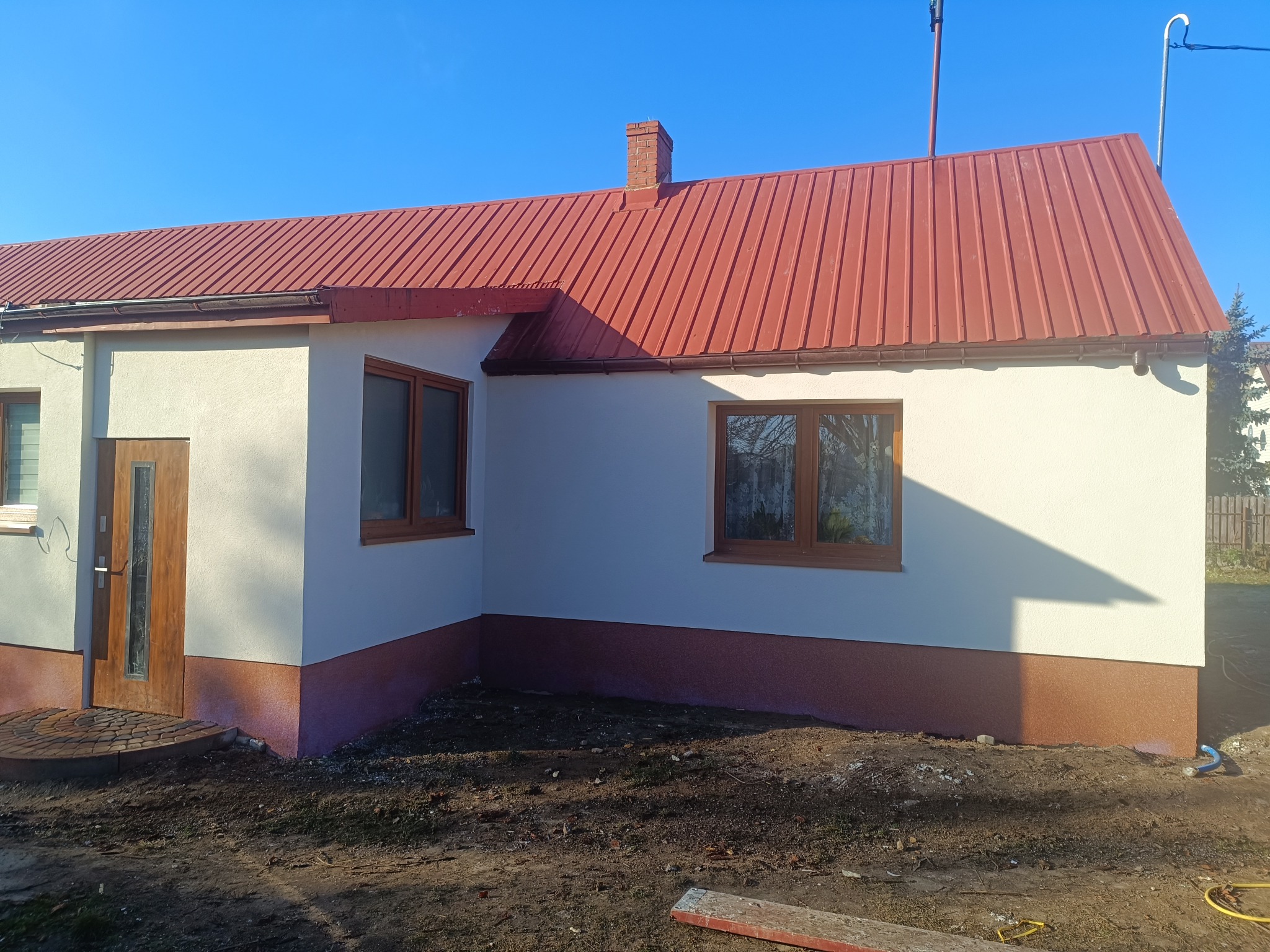 Odnowiony dom jednorodzinny z czerwoną blachodachówką i jasną elewacją, widoczny z boku w słoneczny dzień. Dolna część elewacji w kolorze brązowym.