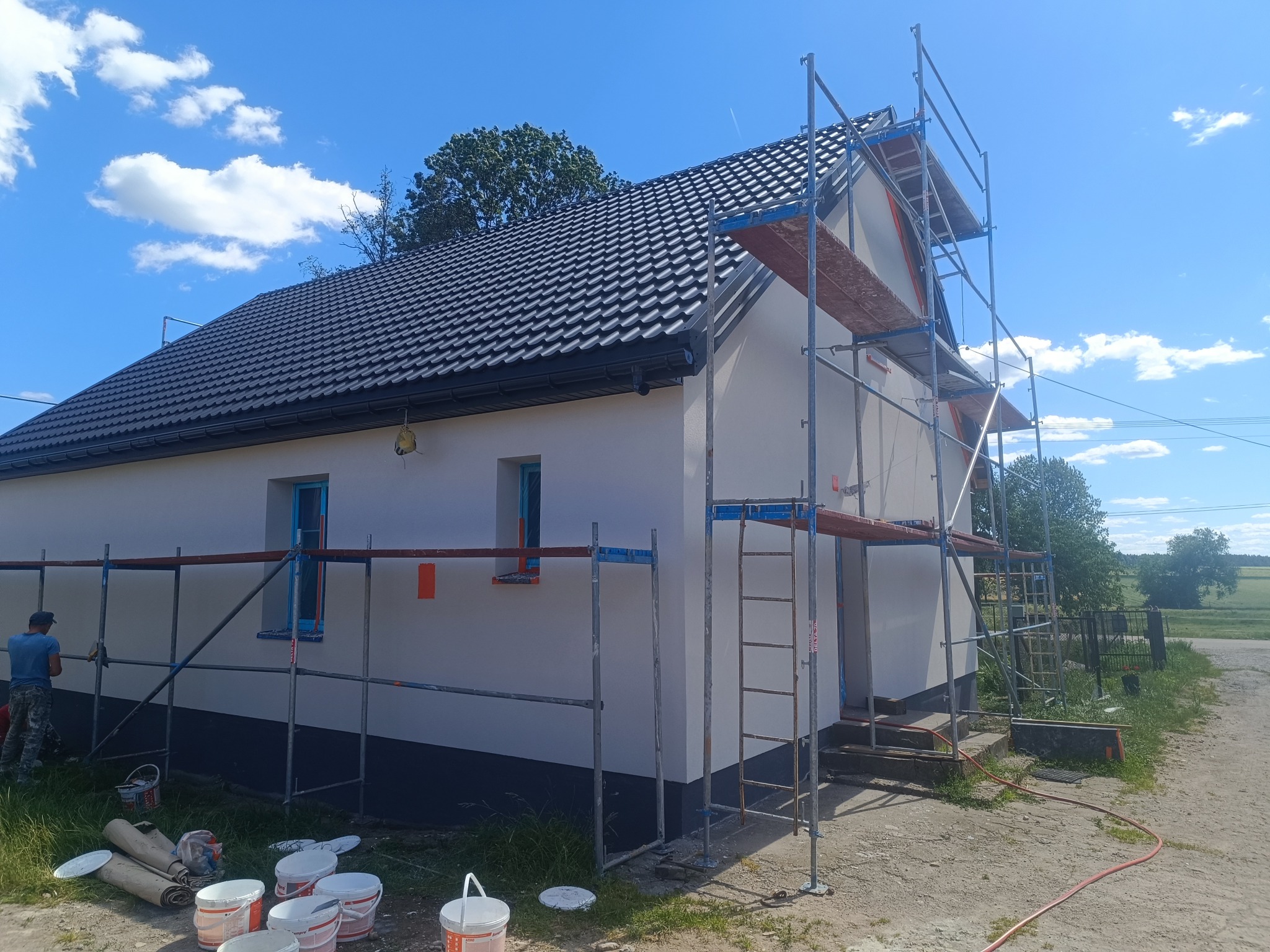 Dom jednorodzinny w trakcie prac elewacyjnych, otoczony rusztowaniem. Widoczne farby i narzędzia, błękitne ramy okienne, czarny dach i fragment sylwetki pracownika.