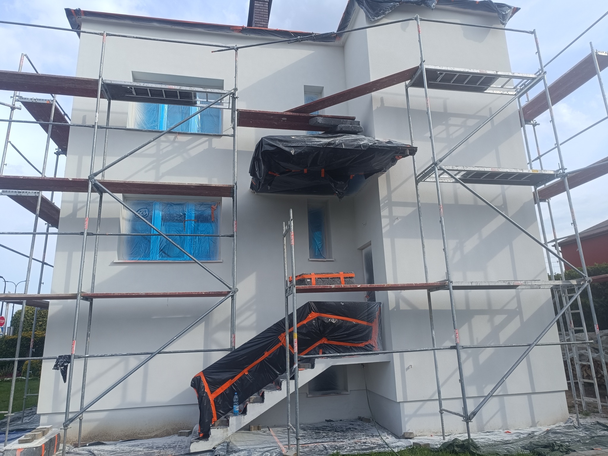 Biała elewacja domu w trakcie prac wykończeniowych. Widoczne rusztowania, okna zabezpieczone folią, schody zakryte plandeką. Świeżo pomalowana fasada.