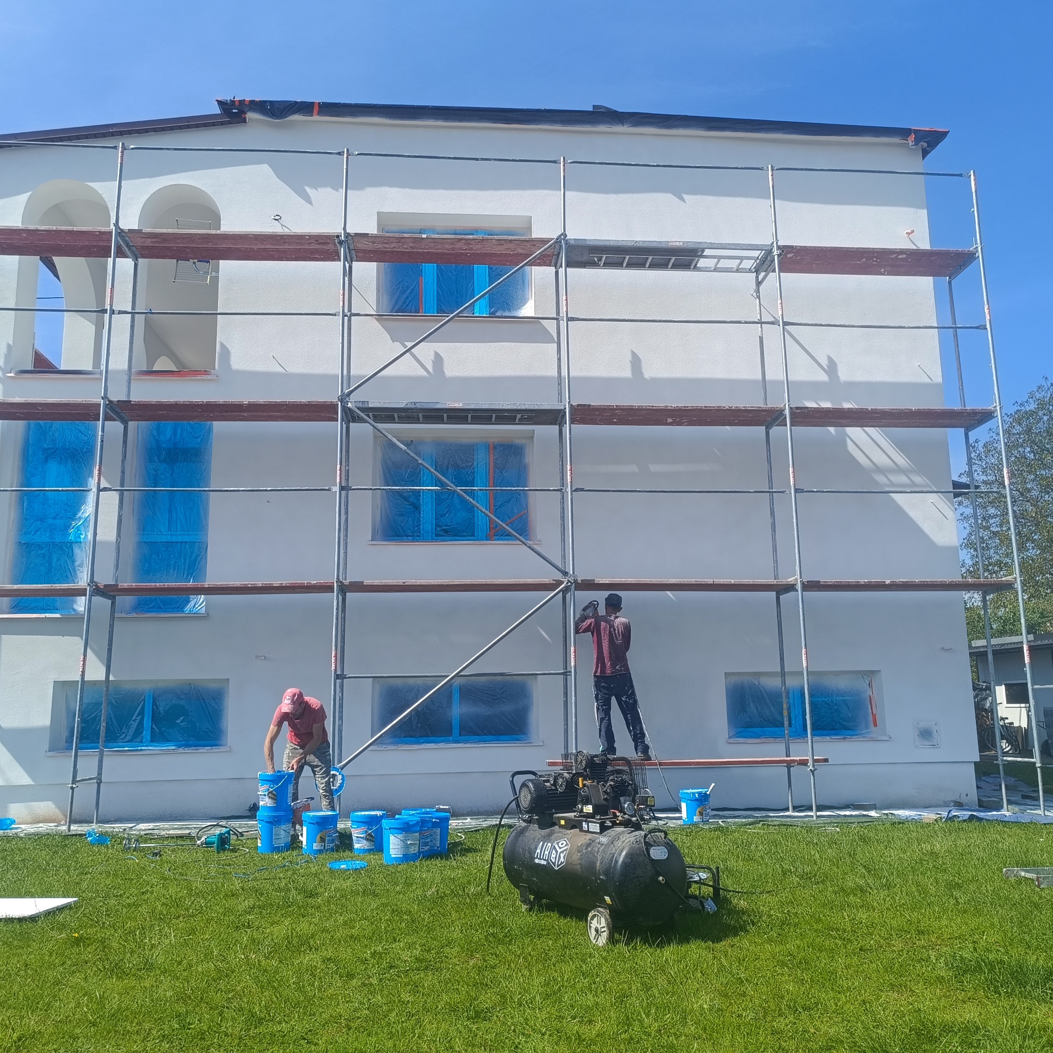 Dwóch pracowników na rusztowaniu maluje białą elewację budynku. Okna zabezpieczone folią, na trawie kompresor i wiadra z farbą. Słoneczny dzień.