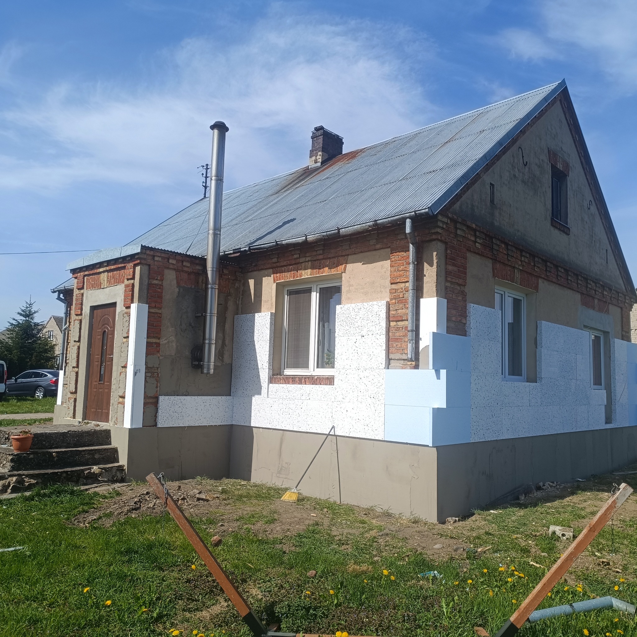 Dom jednorodzinny w trakcie prac związanych z ociepleniem elewacji. Widoczne fragmenty styropianu na ścianach i przygotowane podłoże. Na pierwszym planie trawa i narzędzia.
