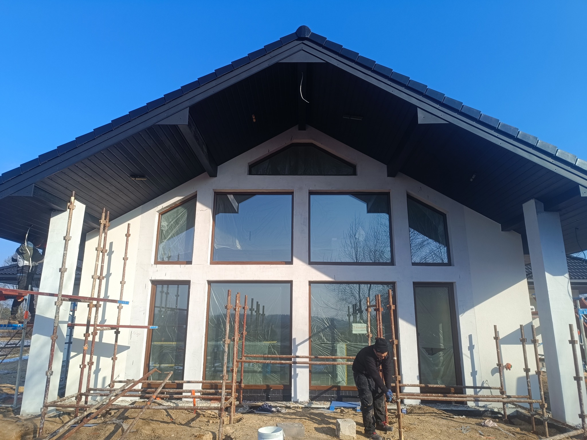 Prace na elewacji domu z dużymi oknami. Widoczne rusztowania i pracownik w trakcie montażu. Świeżo otynkowana fasada w jasnym kolorze, czarny dach i stolarka okienna.