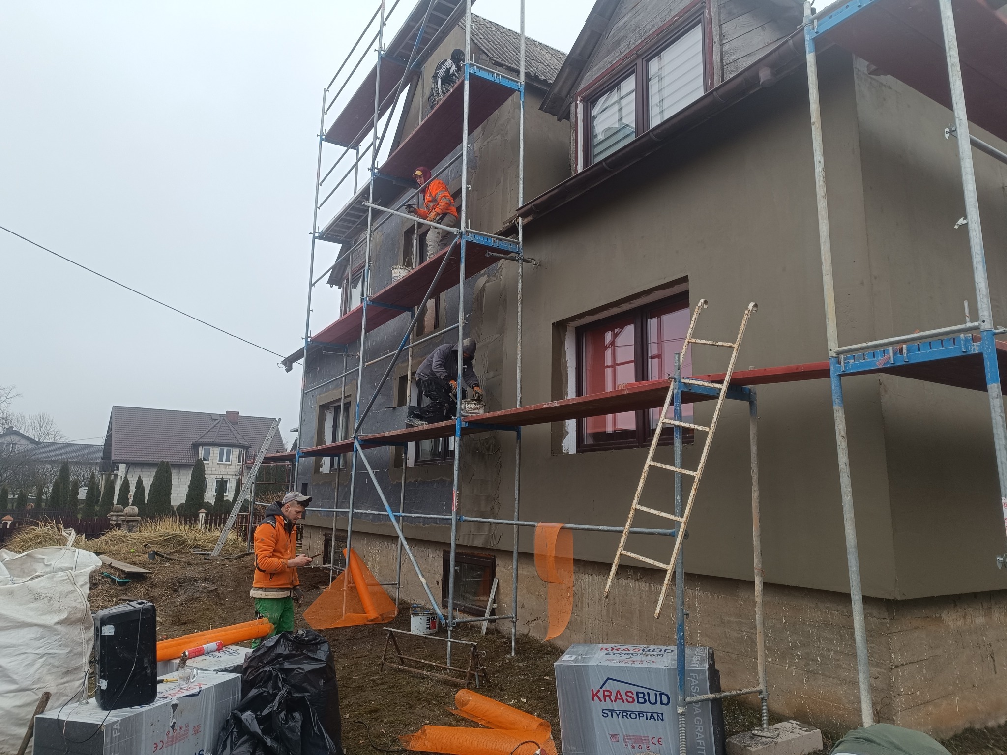 Ekipa na rusztowaniu podczas prac elewacyjnych domu. Widoczne materiały izolacyjne i narzędzia. Częściowo otynkowana ściana, drabina oparta o budynek.