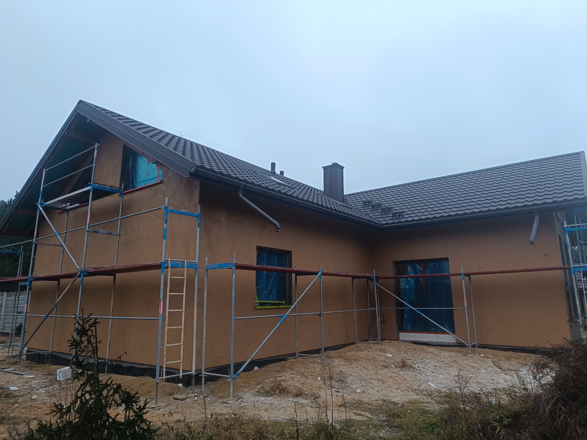 Dom jednorodzinny w trakcie prac elewacyjnych, z widocznymi rusztowaniami i zabezpieczonymi oknami. Ściany pokryte brązowym tynkiem, dach z ciemnej dachówki.