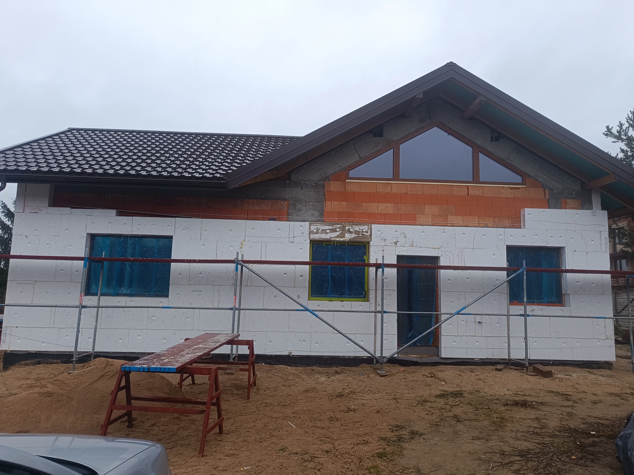 Dom w trakcie ocieplania elewacji styropianem, widoczne rusztowania i zabezpieczone okna folią. Część ściany z cegły i okno trójkątne na poddaszu. Dach pokryty brązową dachówką.