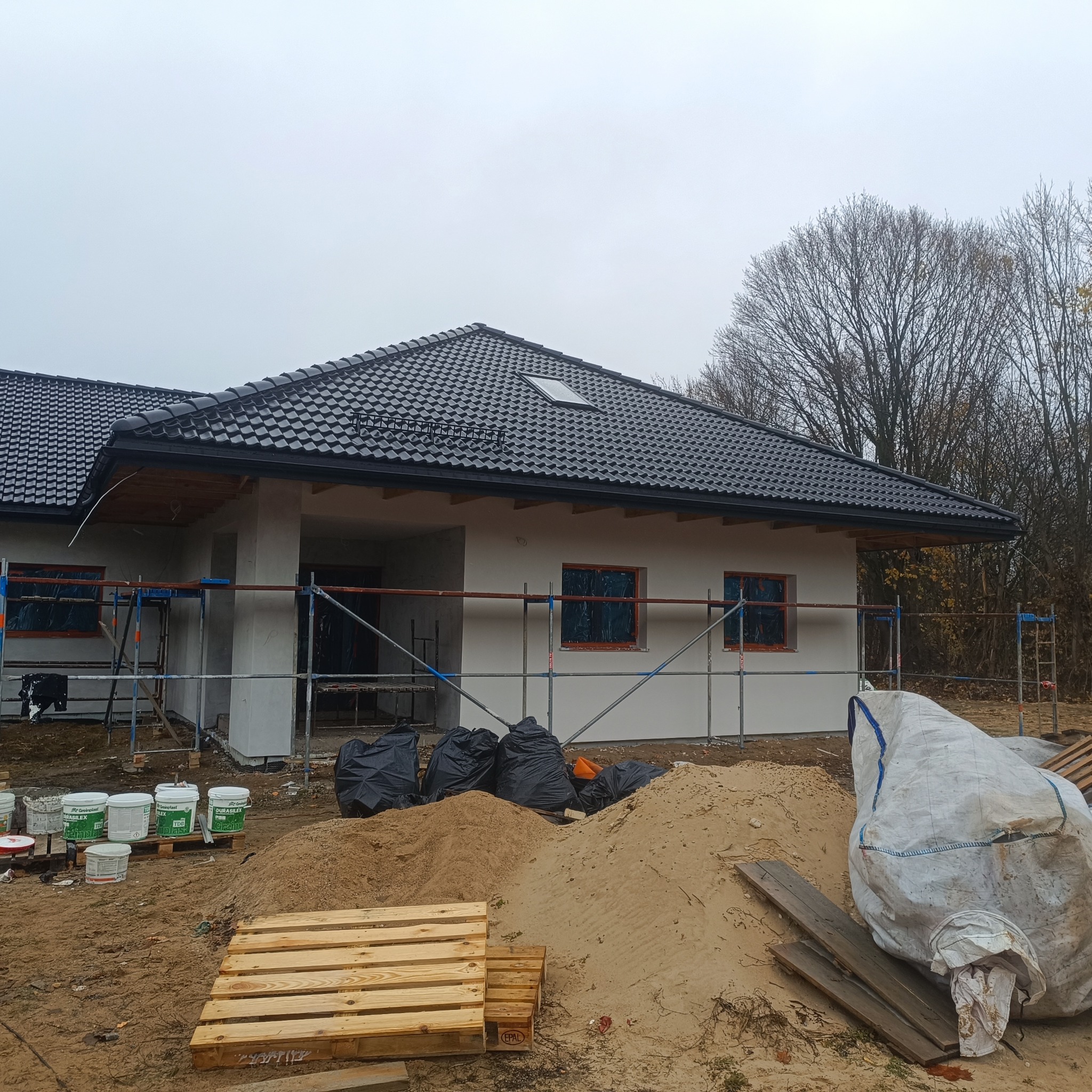 Dom jednorodzinny w trakcie prac wykończeniowych elewacji. Widoczne rusztowania, worki z odpadami i puste wiadra po farbach. Dach pokryty czarną dachówką.