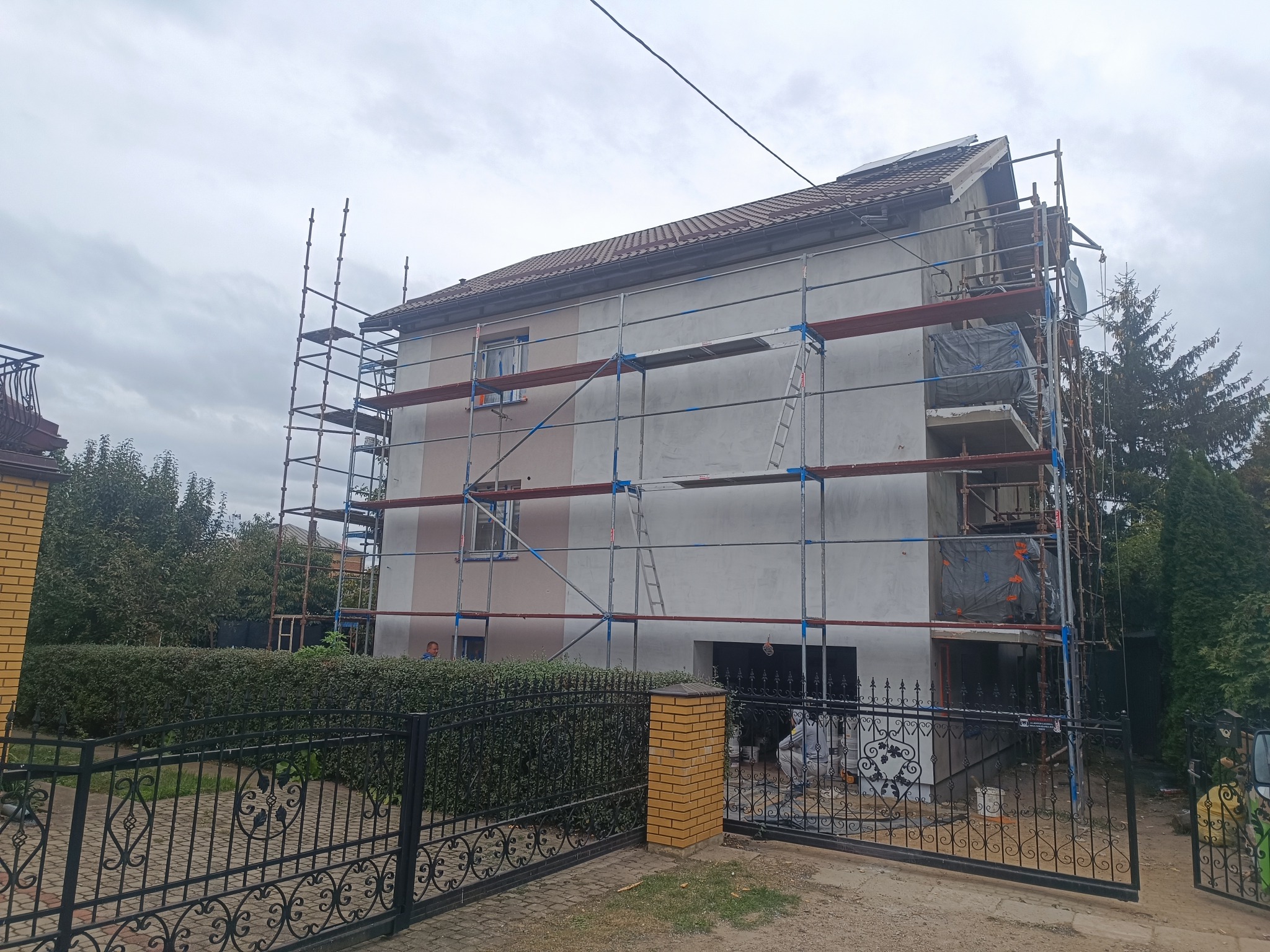 Dom w trakcie prac elewacyjnych, otoczony rusztowaniem. Widoczne fragmenty ocieplenia i przygotowania do nałożenia warstwy wykończeniowej. Prace w toku.