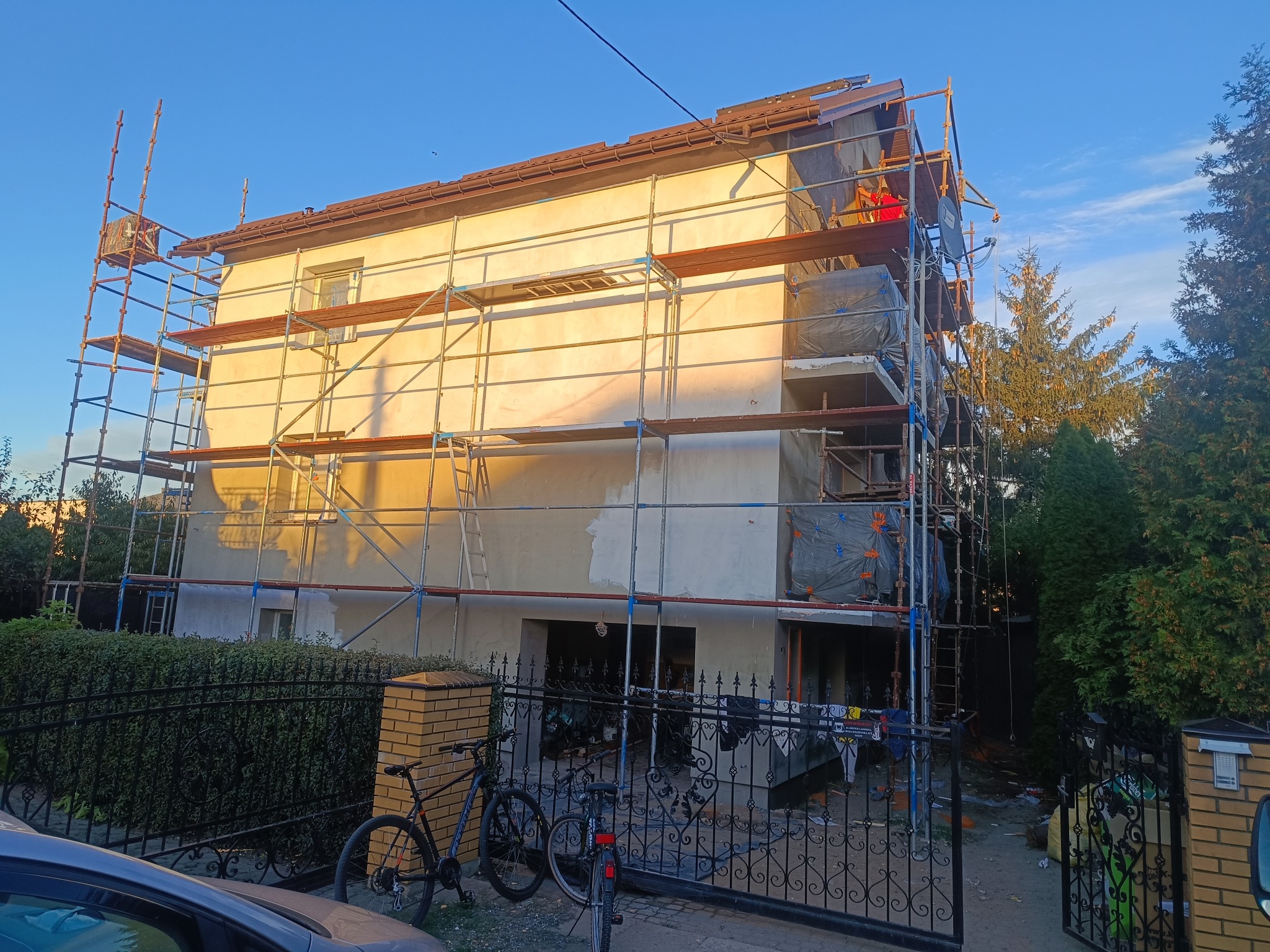 Dwupiętrowy dom w trakcie prac elewacyjnych, otoczony rusztowaniami. Widoczne fragmenty otynkowanej ściany i pracownik w czerwonej koszulce na górze rusztowania.