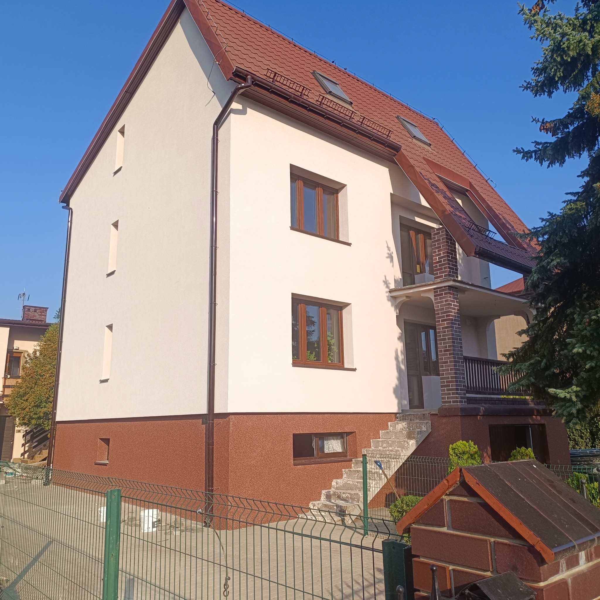 Dwukondygnacyjny dom z beżową elewacją i brązowym cokołem, widoczny zza zielonego ogrodzenia. Dach z brązowej dachówki, okna w drewnianych ramach.