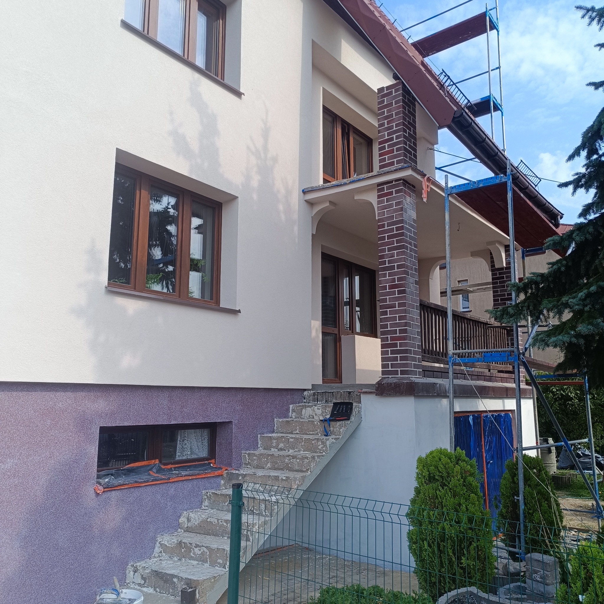 Elewacja domu po odświeżeniu, z elementami dekoracyjnymi z cegły. Widoczne rusztowania, sugerujące trwające prace wykończeniowe. Dolna część ściany w innym kolorze.