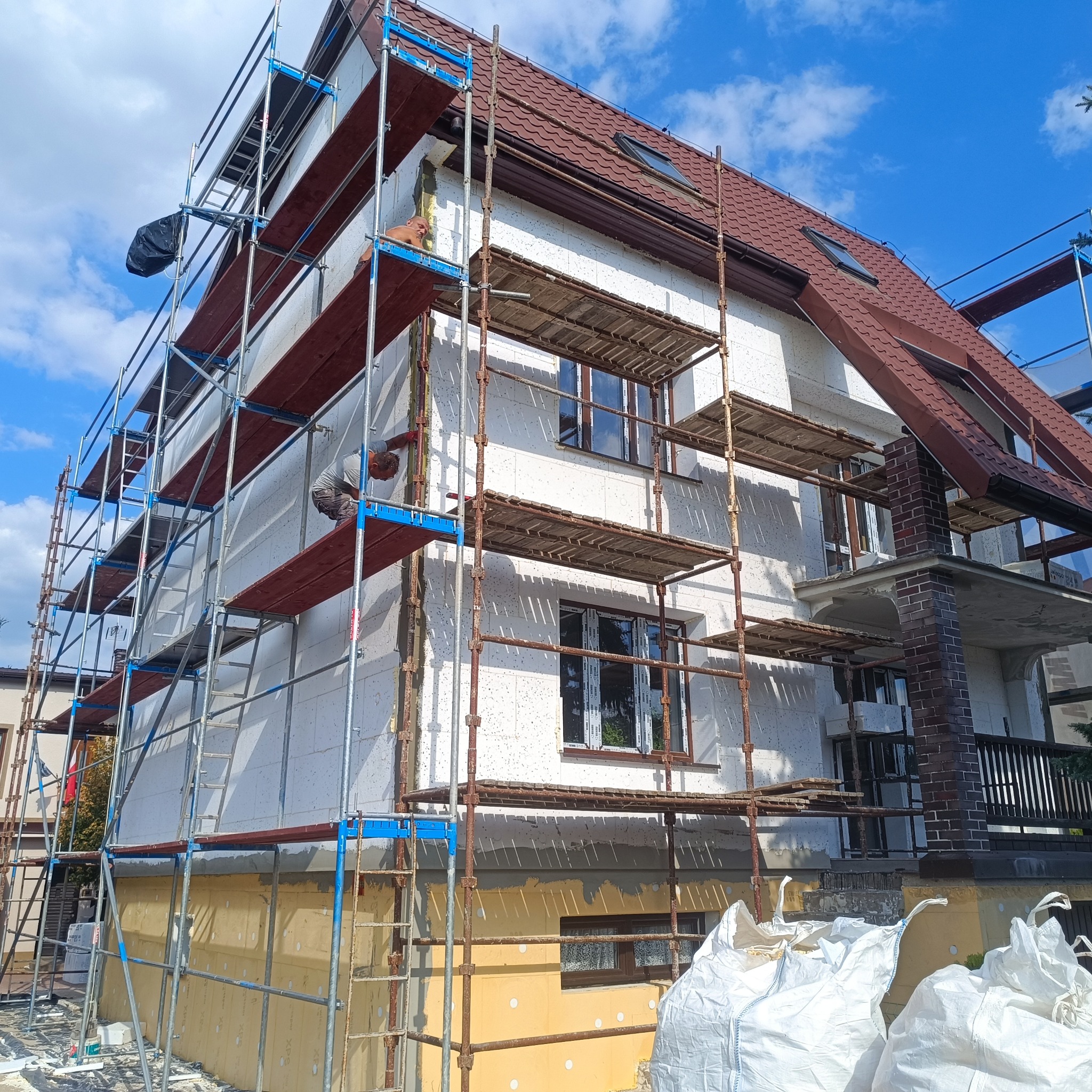 Dom w trakcie ocieplania, widoczne rusztowania, styropian na elewacji i pracownik. Częściowo ocieplona dolna partia budynku. Słoneczny dzień, błękitne niebo.