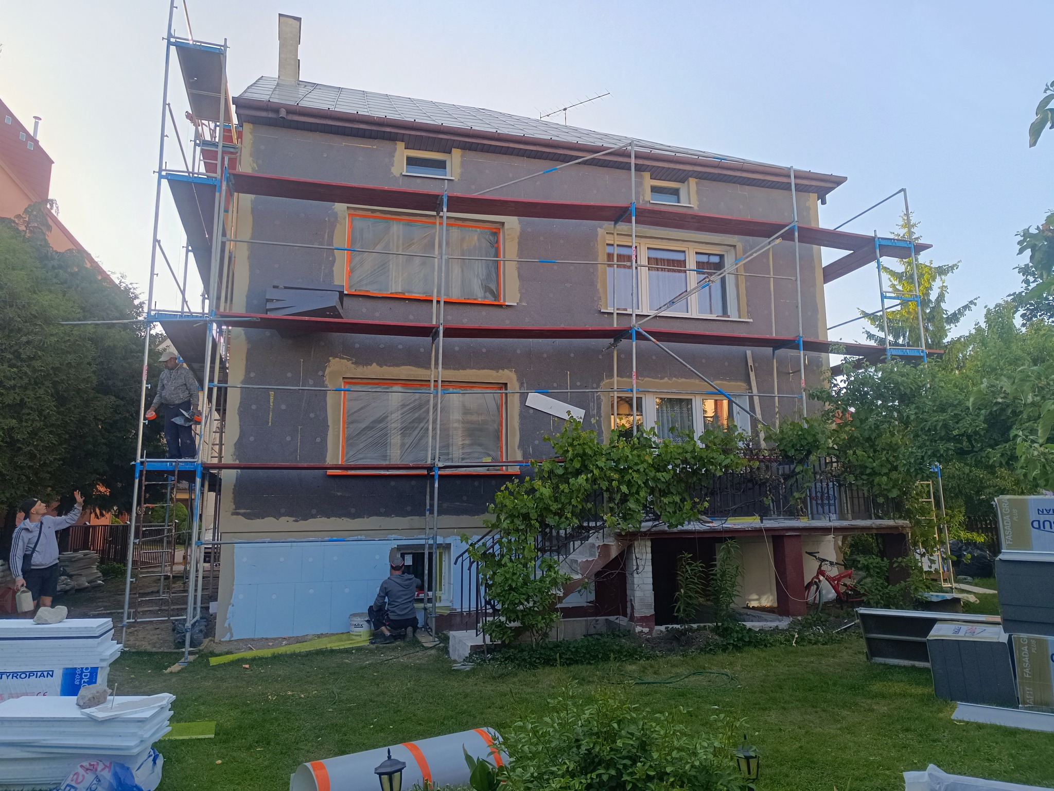 Dwupiętrowy dom w trakcie ocieplania elewacji, widoczne rusztowania, pracownicy i materiały izolacyjne. Okna zabezpieczone folią. Część elewacji pokryta styropianem.