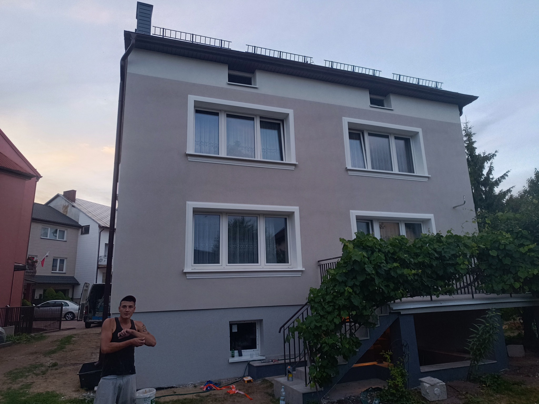 Dwukondygnacyjny dom po renowacji elewacji w odcieniach szarości i bieli, z widocznymi oknami w białych ramach i elementami zieleni. Mężczyzna stoi przed budynkiem.
