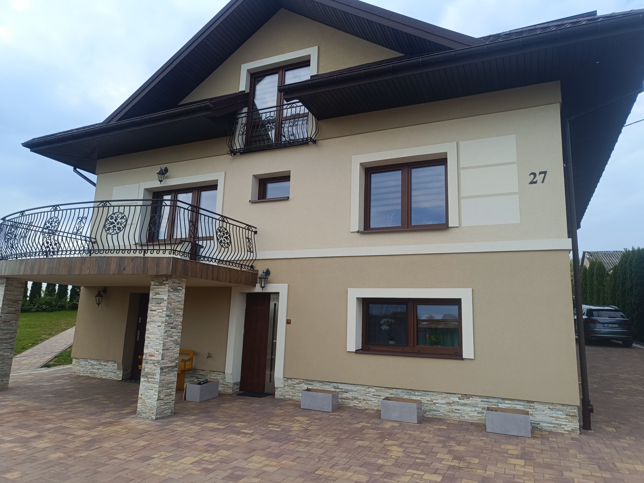 Dwupiętrowy dom z beżową elewacją, balkonami z ozdobnymi balustradami i numerem 27. Widoczny fragment podjazdu z kostki brukowej i samochód w tle.