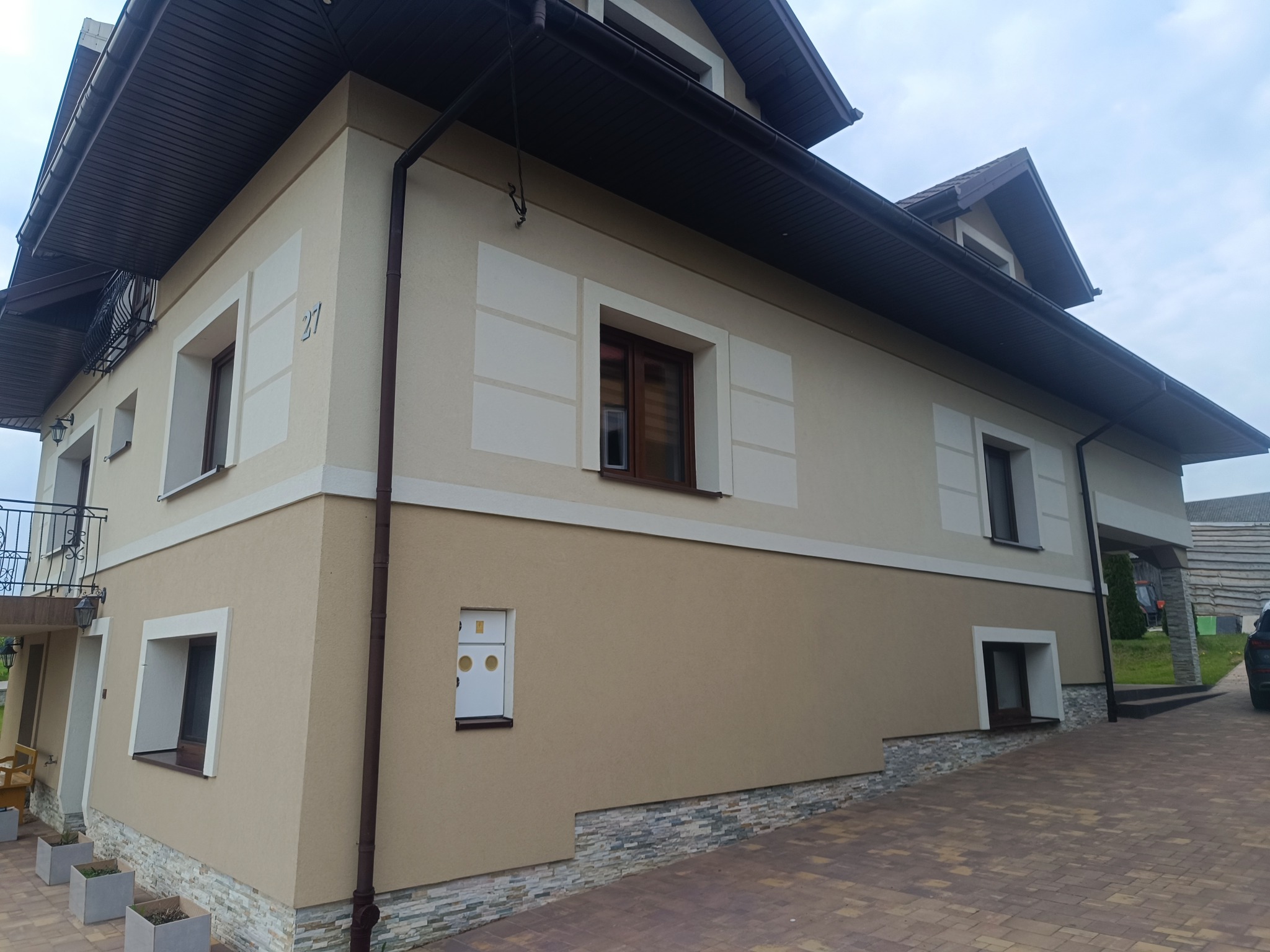 Dwukondygnacyjny dom z beżową elewacją, białymi akcentami wokół okien i ciemnym dachem. Widoczny fragment brukowanego podjazdu i zadbany ogród. Numer domu '27' na ścianie.